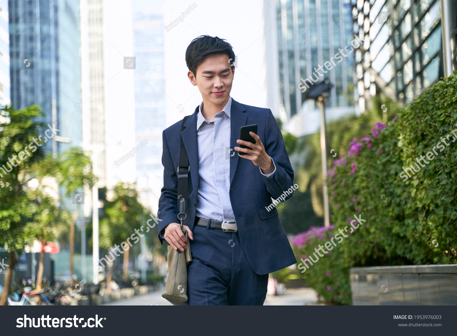 young asian businessman looking at messages on cellphone while walking in the street in downtown of modern city #1953976003