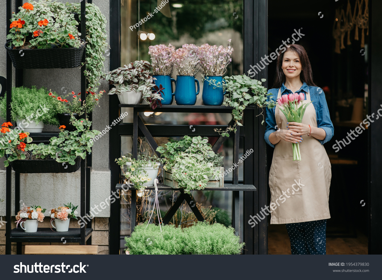 Woman florist reopening shop after covid-19 pandemic, delivery service for clients. Happy young female in apron holds bouquet of tulips near front door of store with vases and pots of diverse flowers #1954379830
