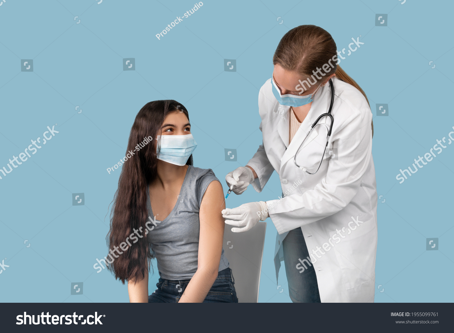 Medical doctor or nurse giving coronavirus vaccine shot to Indian teen girl over blue studio background. Female teenager in face mask getting covid-19 vaccine, participating in immunization campaign #1955099761