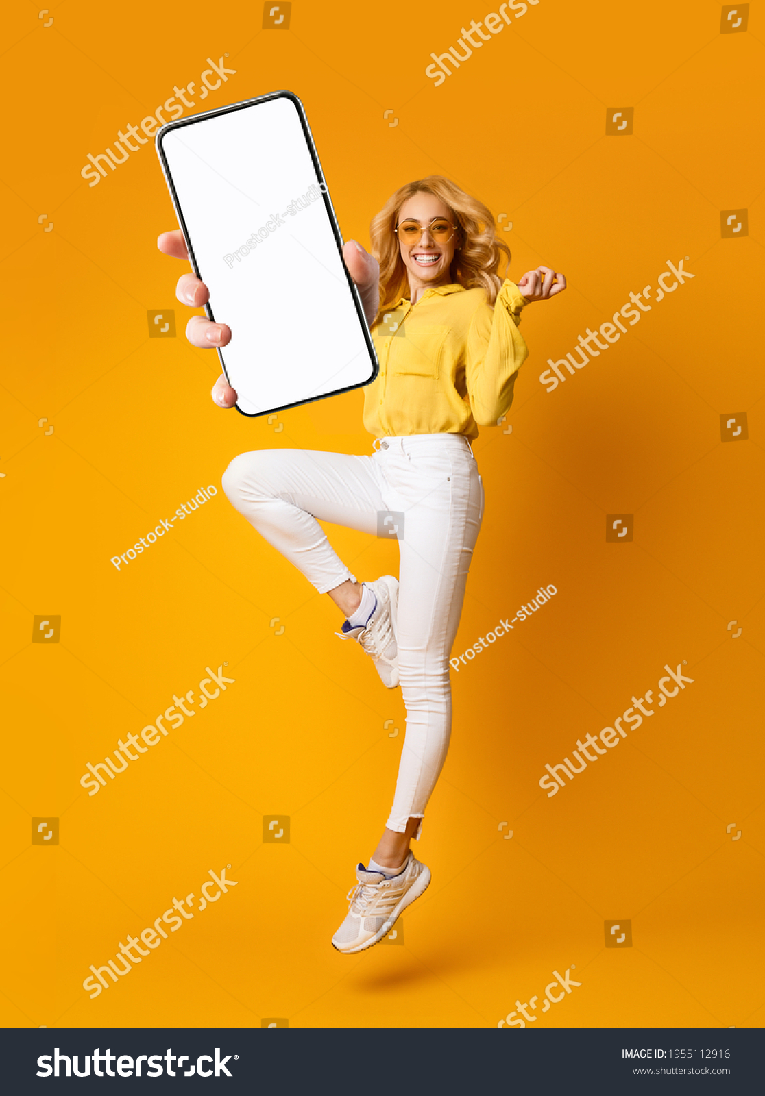 Cheerful blonde woman jumping up and showing newest smartphone with empty screen, enjoying new application for mobile device. Orange studio background, creative collage with mockup #1955112916