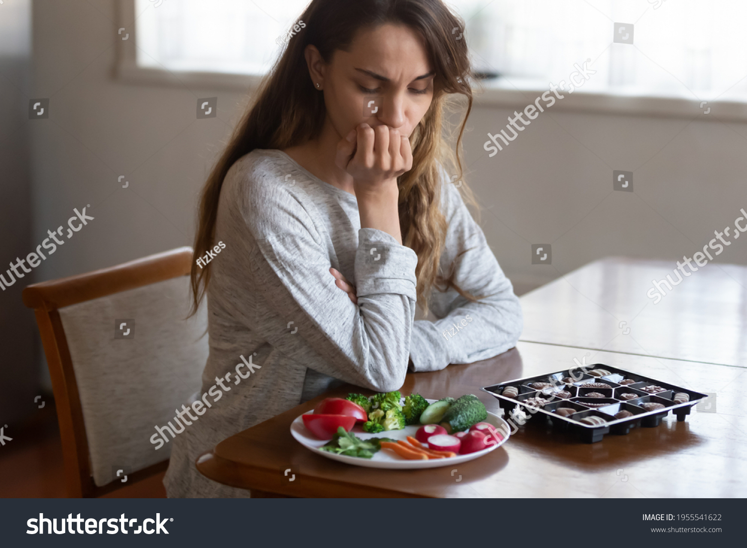 Unhappy young Latin woman look at chocolates and vegetables face temptation suffer from eating disorder. Millennial female think of healthy food choice, diet. Wellbeing, weight loss concept. #1955541622