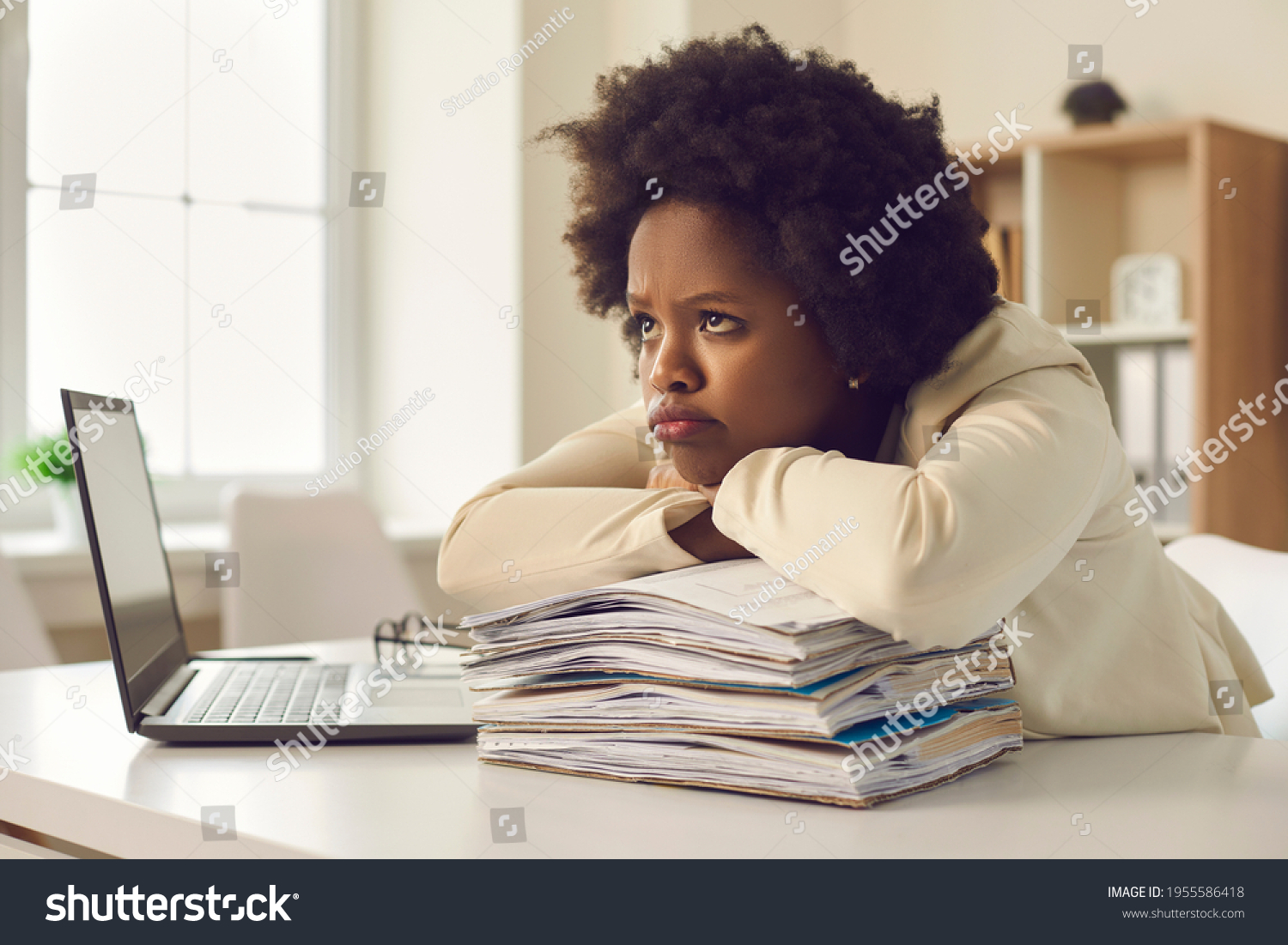 Bored unsatisfied young woman waiting for working day to end. Tired employee dissatisfied she has to work extra hours and do all paperwork sitting at office desk pouting lips with sad face expression #1955586418