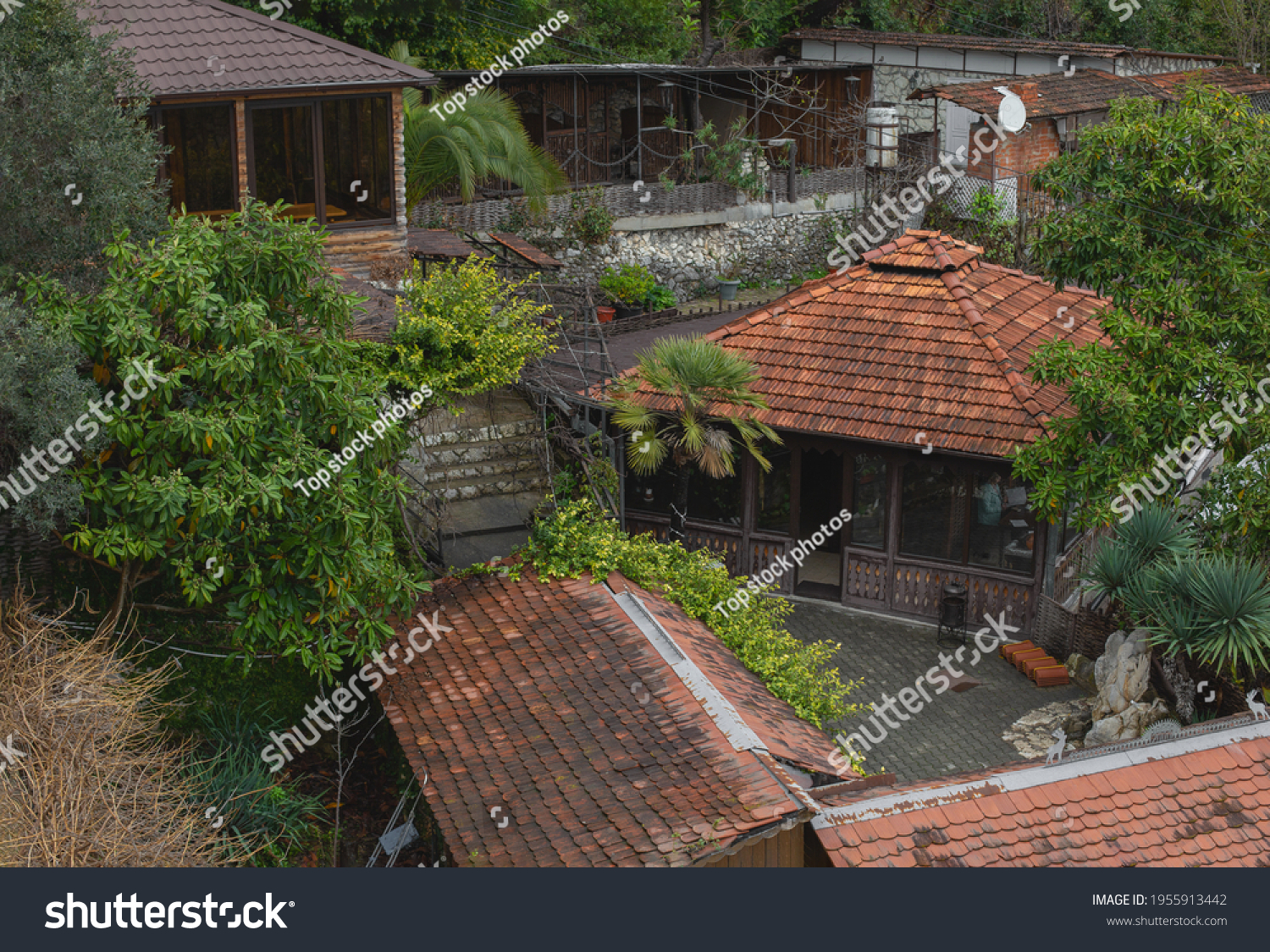 View of the old houses with red tiled roofs and green trees. Old garden with palm and other plants. Abandoned buildings. Shack. Hovel. #1955913442
