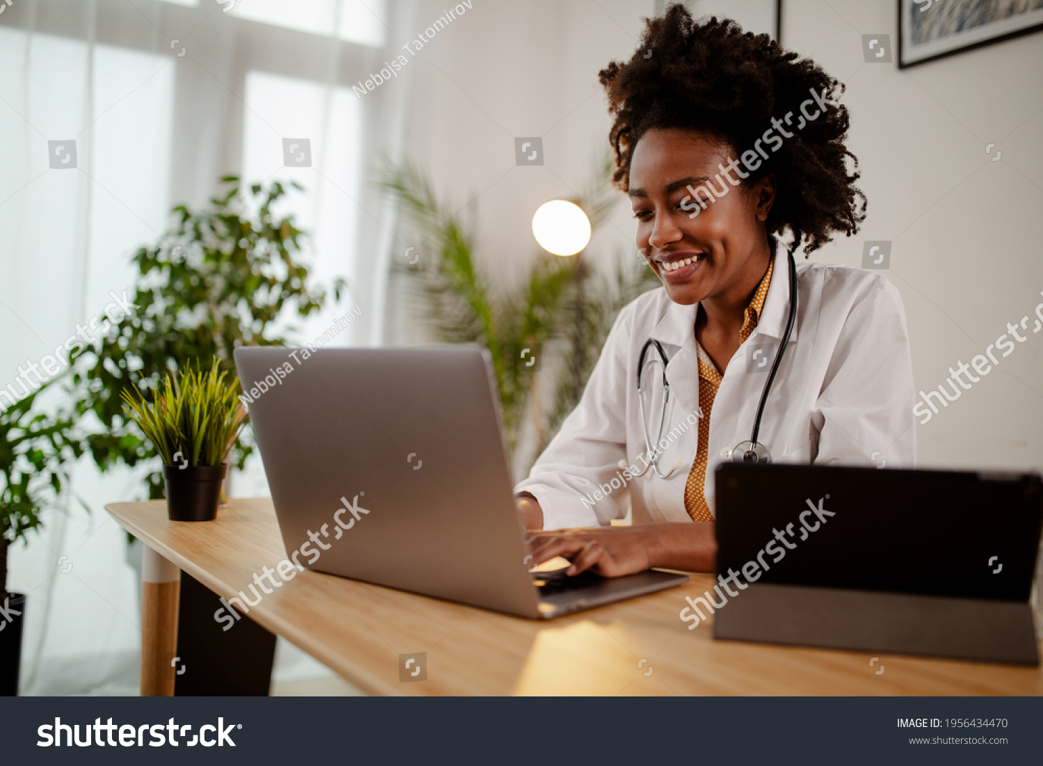 Successful young Afro American female doctor in white medical uniform and stethoscope busy working on laptop in modern hospital, smiling woman nurse or GP typing on computer consult patient online. #1956434470