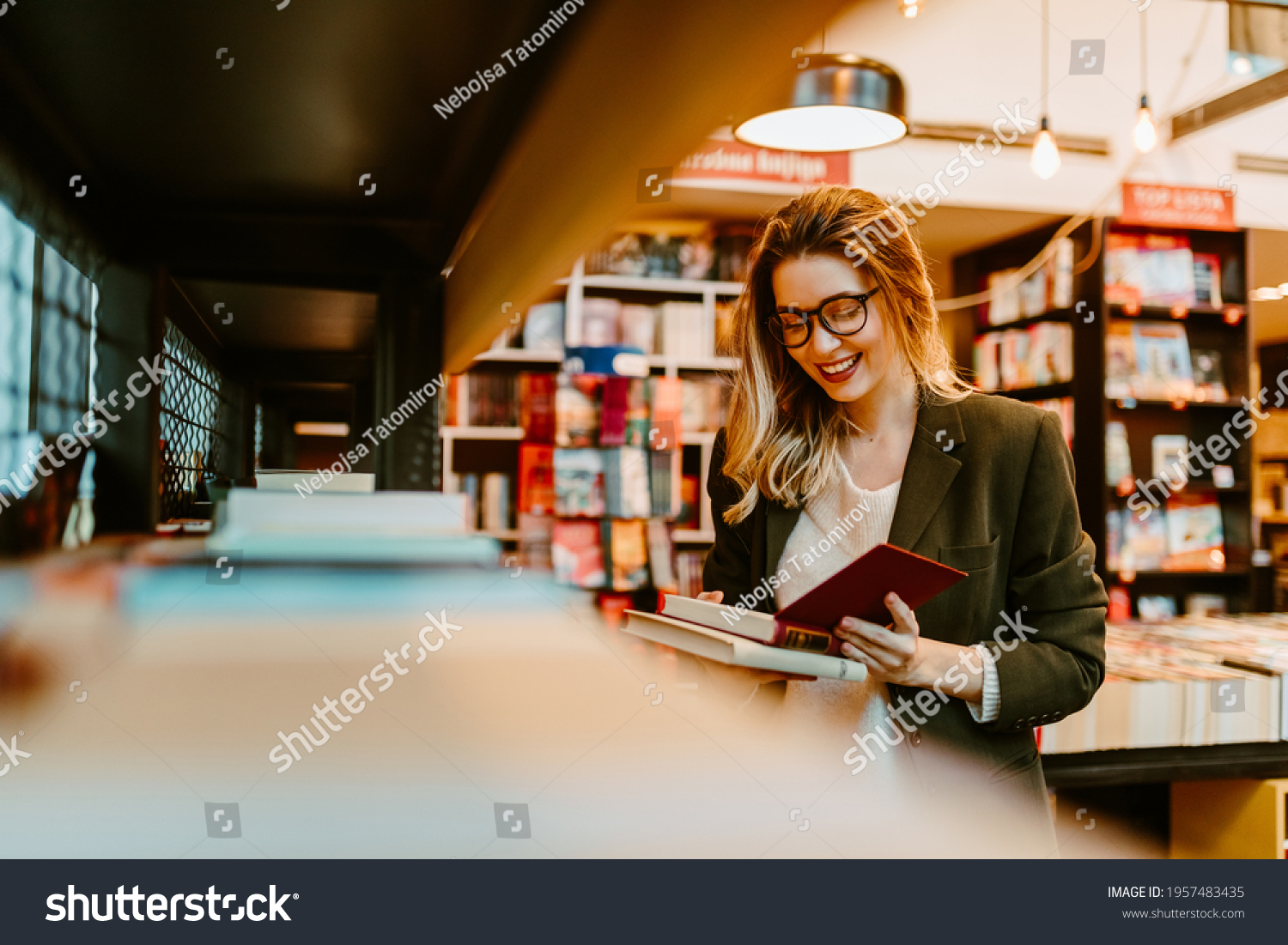 Photo of young woman reading impressum for book that she just grabbed from book shelf. Young woman is spending her free time at bookstore. #1957483435