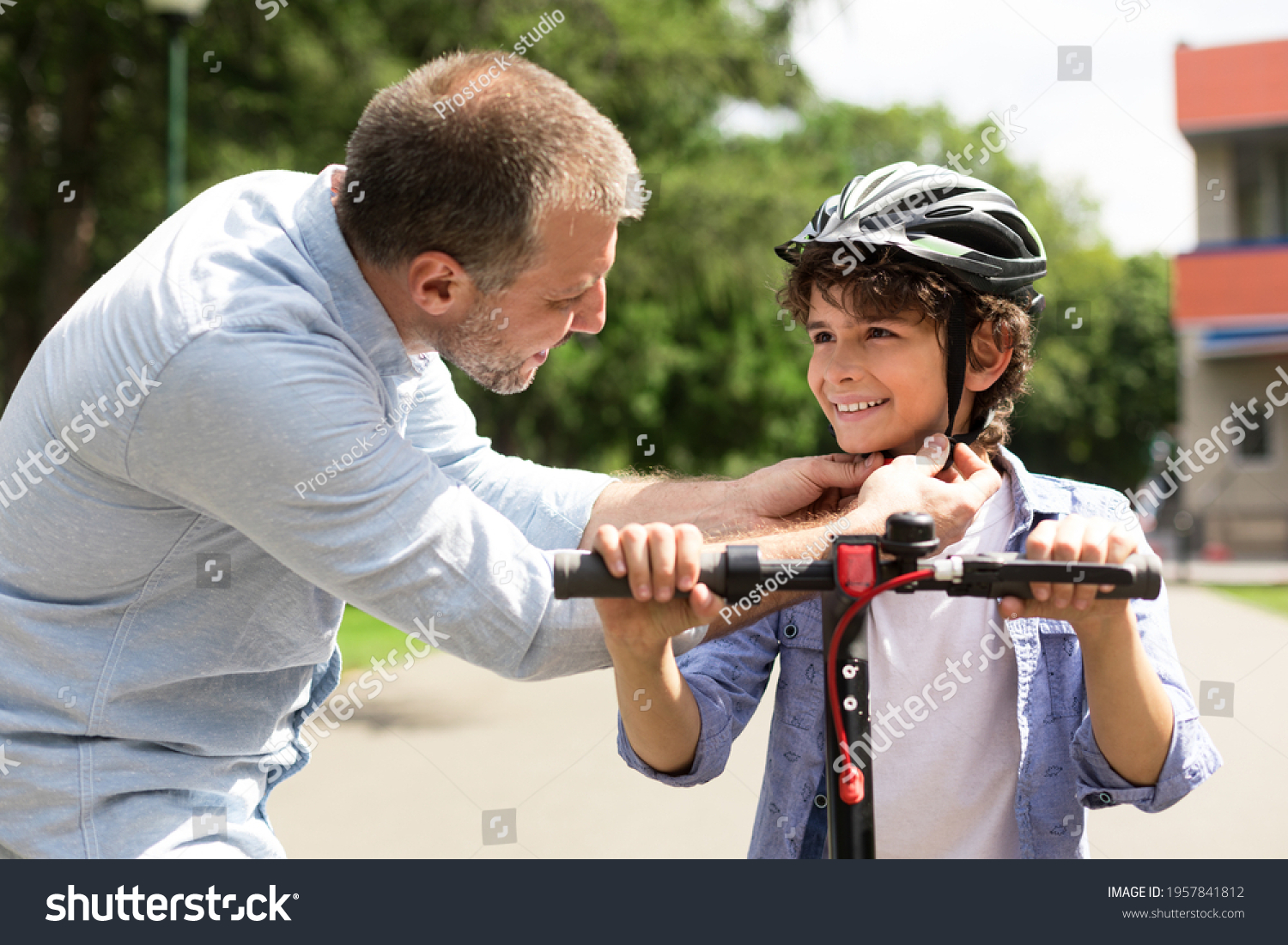 Safety Concept. Close up portrait of smiling adult father putting protection helmet on little boy at park, teaching his brave son to ride electric motorized scooter. Ourdoor Weekend Activity #1957841812