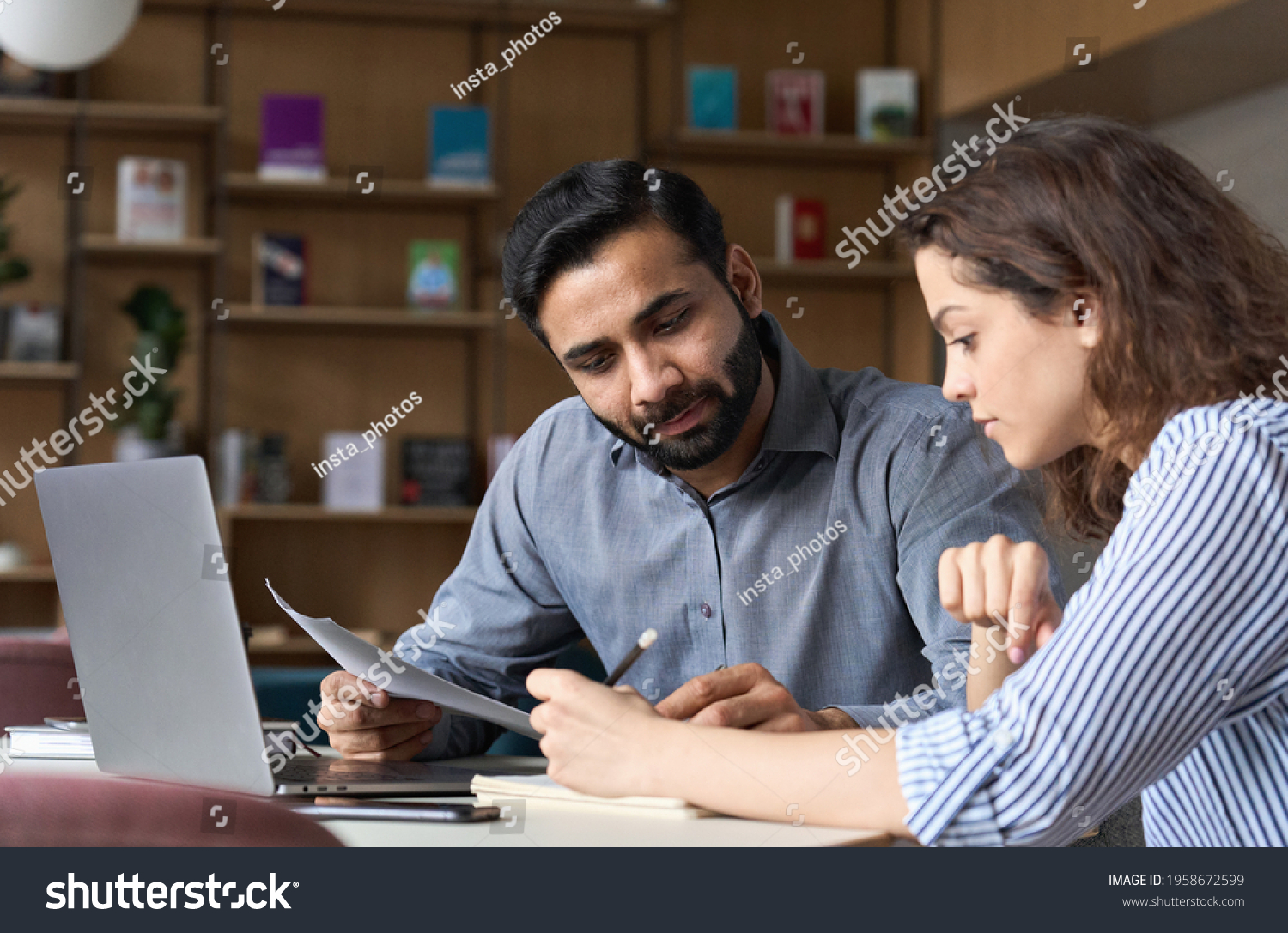 Two multiethnic professionals colleagues working together with laptop and papers in office. Indian male mentor and latin female young professional sitting in creative office space. #1958672599