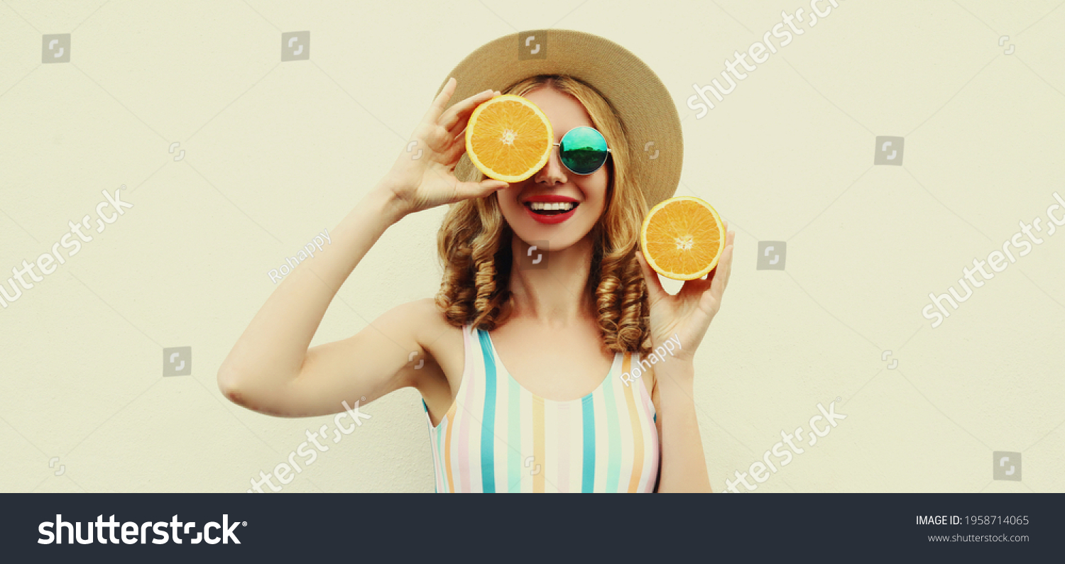 Summer positive portrait of cheerful smiling young woman covering her eyes with slices of orange wearing a straw hat on a white background #1958714065
