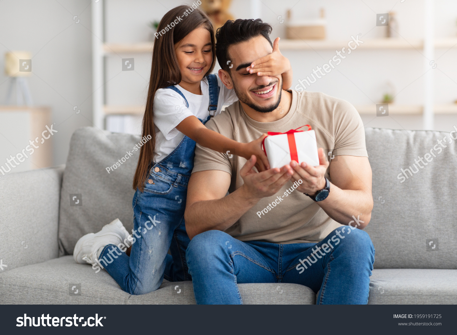 Portrait of cute little girl holding and giving wrapped gift box, making surprise for her excited dad, covering his eyes, greeting young man with father's day or birthday, celebrating at home #1959191725