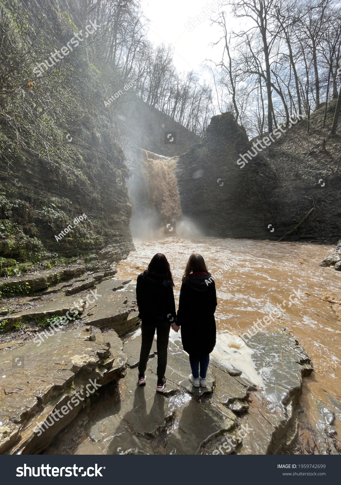 Rear view of two women looking on powerful muddy waterfall. Tourists standing on rocks in mountainous terrain and enjoying beautiful view of cataract #1959742699