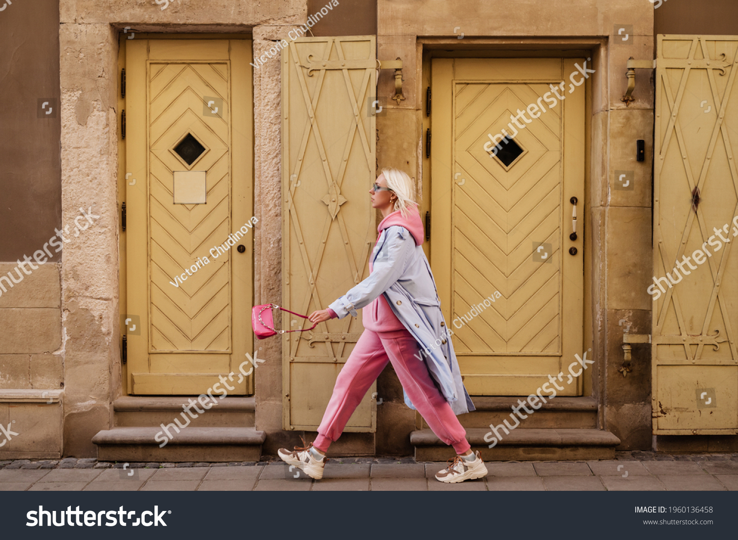 Street style, fashion: woman wearing  trendy sport chic outfit walking in street of European city. Blue trench coat, sunglasses, pink hoodie, trousers, sneakers, mini bag. Copy, empty space for text #1960136458