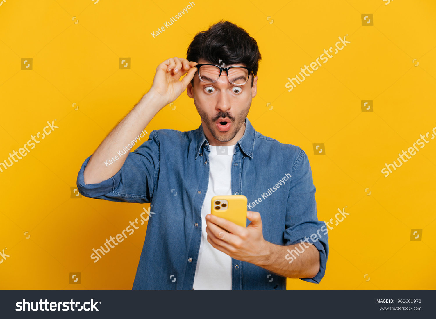 Amazed shocked caucasian guy holding smartphone in his hand, looking at the phone in surprise with his glasses raised, stunned facial expression, stands on isolated orange background #1960660978