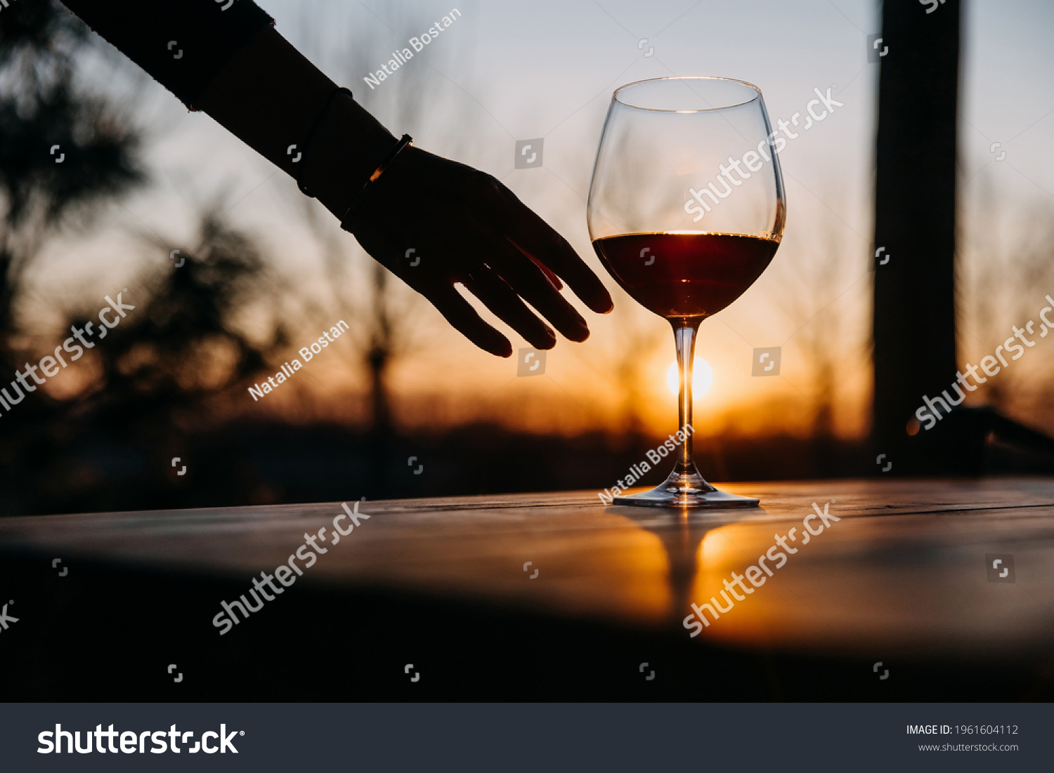 Female hand reaching a glass of red wine on a wooden table, at sunset. #1961604112
