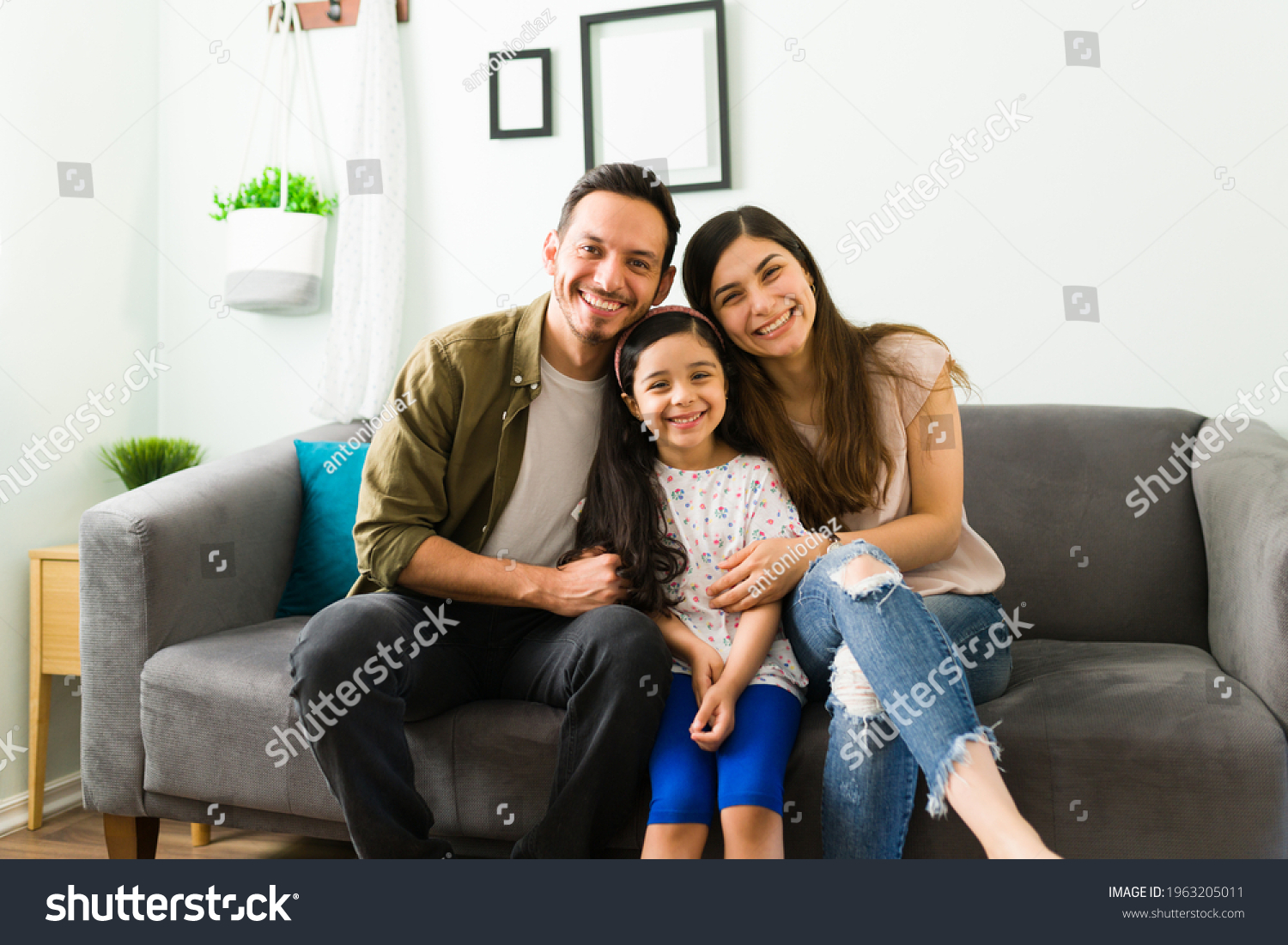 Happy young family of mom, dad and daughter laughing and making eye contact while relaxing together on the living room sofa  #1963205011