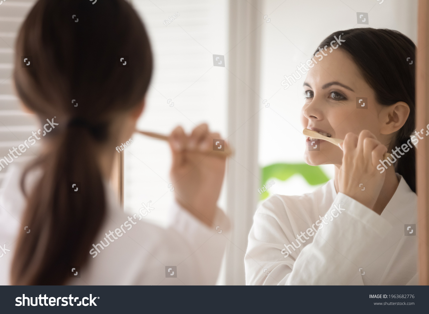 Young woman in bathrobe brushing teeth with wooden bamboo toothbrush. Teen girl choosing eco friendly tools for morning bath routine, oral mouth hygiene, protection from caries. Mirror reflection #1963682776