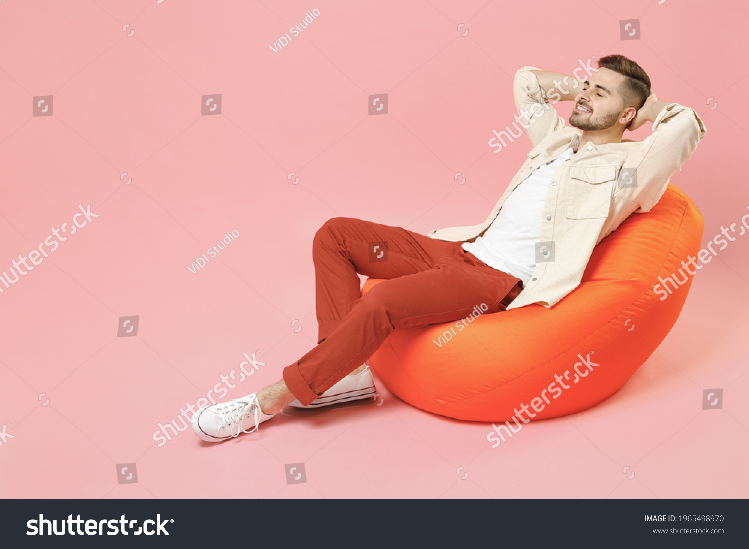 Full length young smiling happy overjoyed joyful fashionable man 20s in jacket white t-shirt sitting in bean bag chair resting hold hand behind neck isolated on pastel pink background studio portrait. #1965498970
