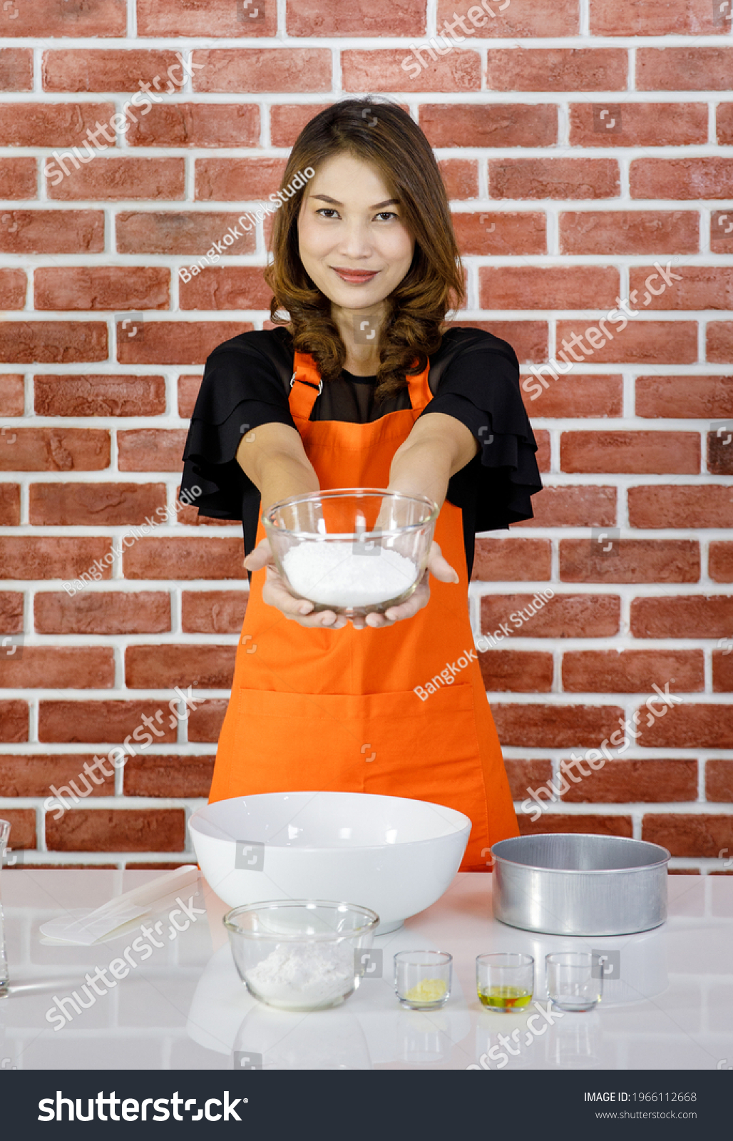 Asian young pastry chef lady in orange apron standing smiling look at camera showing glass bowl of white flour in hand when teaching baking and cooking in front red brick wall kitchen background. #1966112668