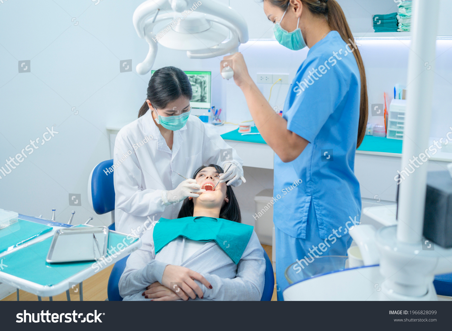 Asian female dentist adjust dental surgical light then starts checking or examining tooth of young girl patient lying on dental chair. Dental assistant support by handing instruments at dental clinic. #1966828099