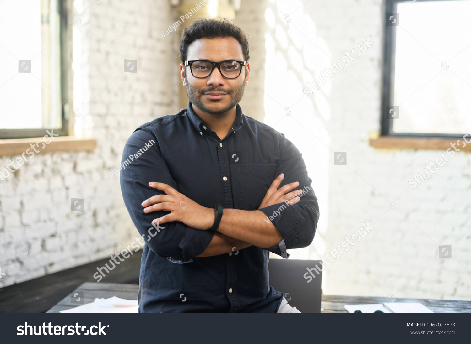 Ambitious hindu man in smart casual shirt stands with arms crossed in contemporary office space and looks at camera, portrait of purposeful indian businessman, ceo indoor. Diversity of office staff #1967097673
