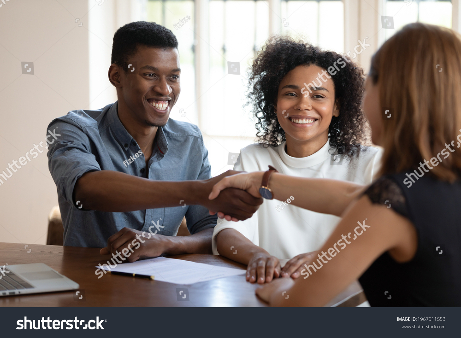 Happy young african american married couple clients shaking hands with financial advisor, discussing contract terms of conditions. Emotional glad millennial diverse spouses celebrating making deal. #1967511553