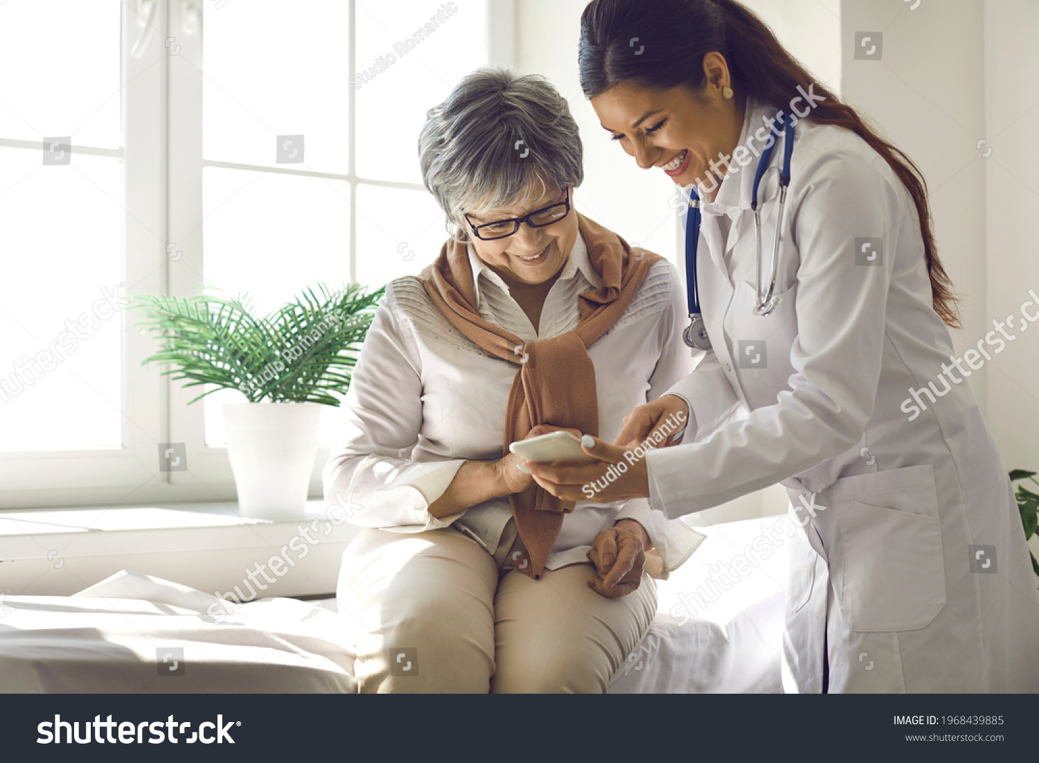 Caring doctor teaches female patient to use mobile healthcare app. Retired lady sitting in hospital exam room looking at cell screen learning to download health tracker for senior citizens. Copy space #1968439885