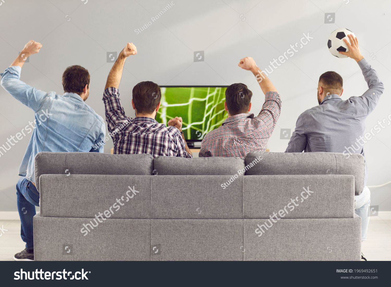 Sofa experts. Back view of male friends gathered at home to watch a football match sitting on the couch in front of the big screen TV. Men actively support their favorite team and comment on the match #1969492651