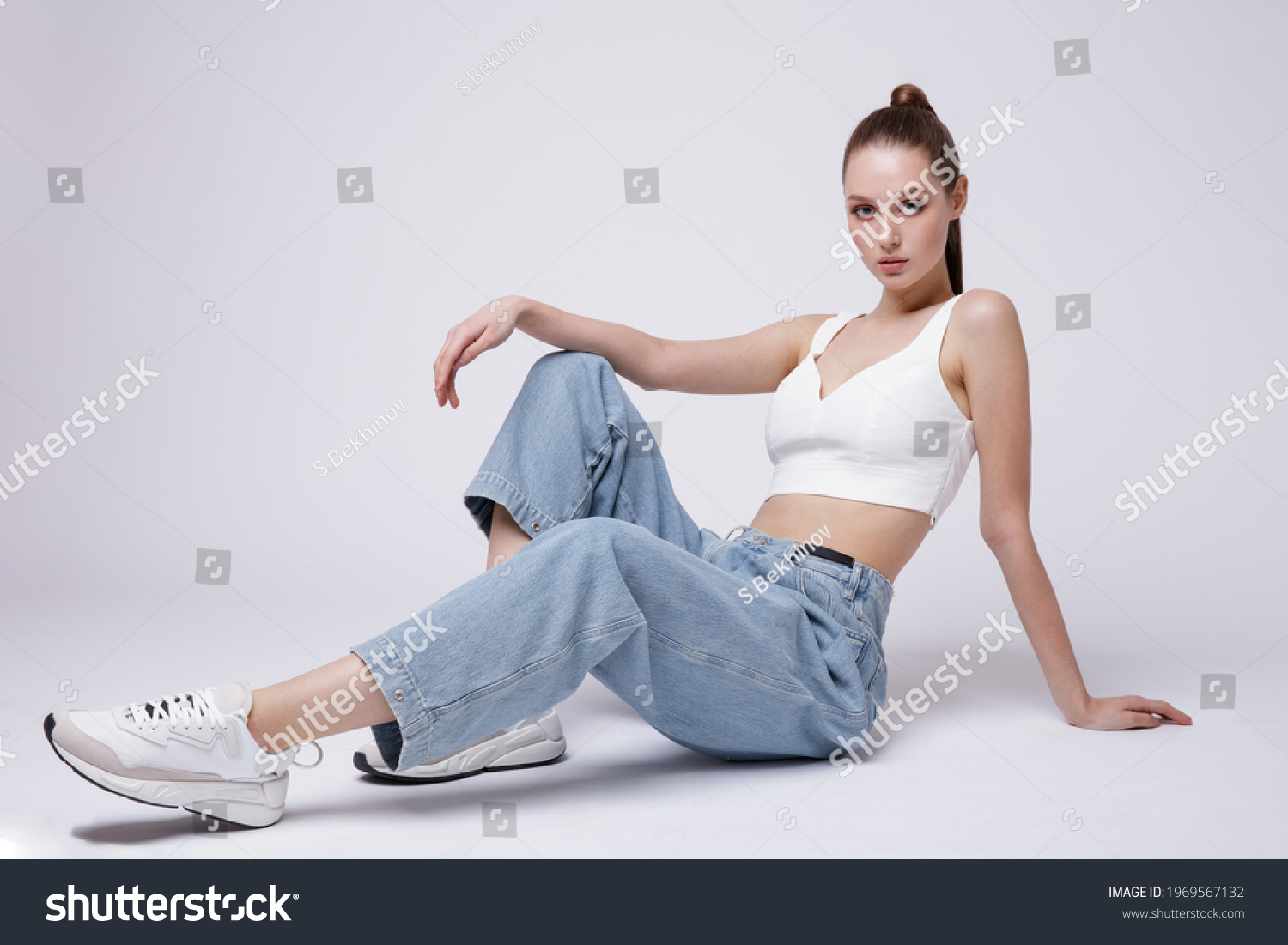 High fashion photo of a beautiful elegant young woman in a pretty white top and sneakers, blue denim jeans posing over white, soft gray background. Studio Shot. The model is sitting #1969567132