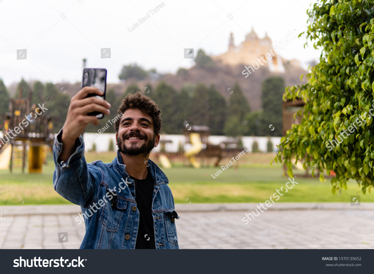 Handsome young man with curly hair and a beard wearing denim and taking a selfie with his smartphone in the pyramid of Cholula, Mexico. #1970139652