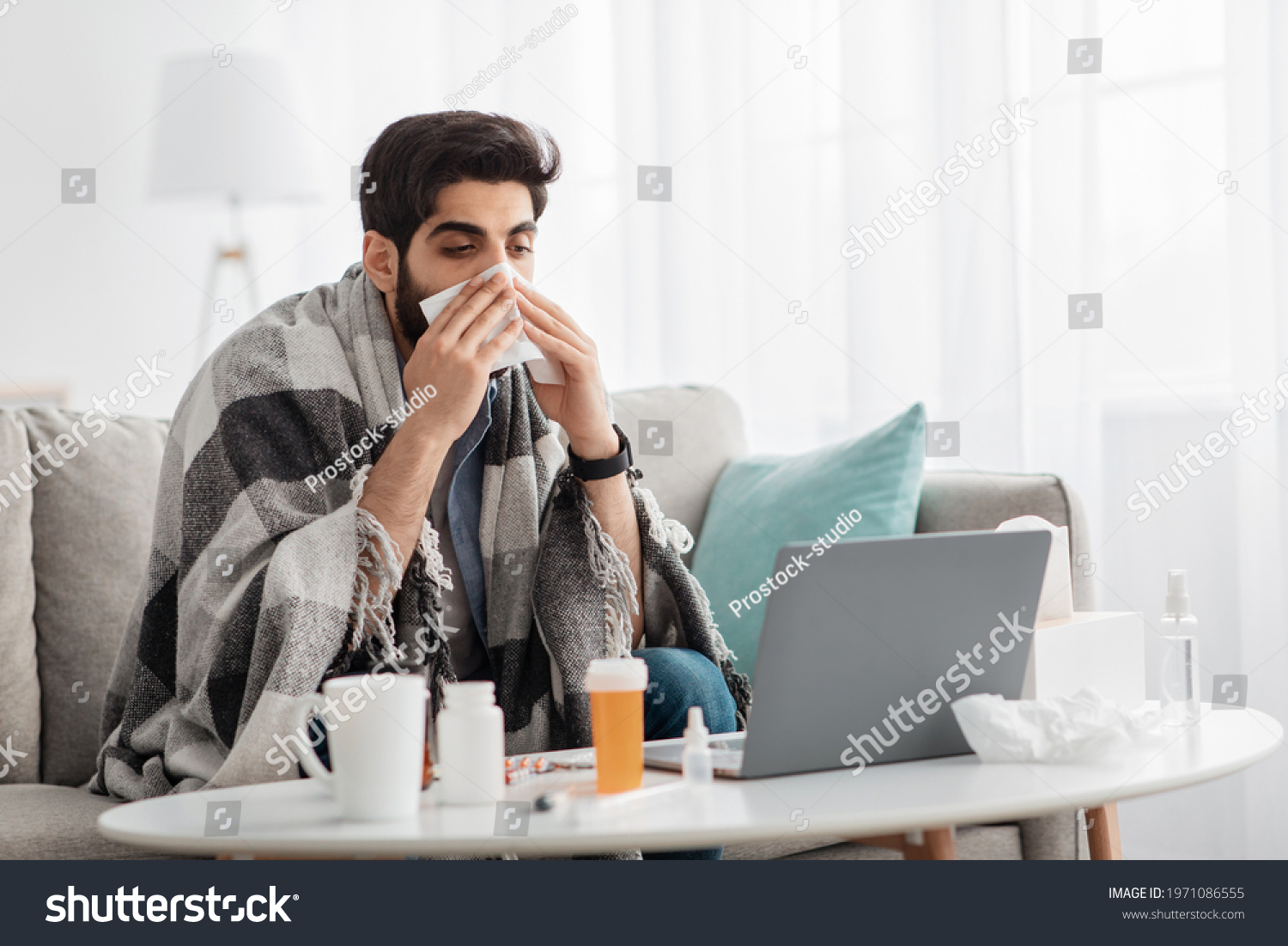 Ill arab man covered in warm blanket looking at laptop screen and sneezing nose in paper tissue, sitting on sofa at home, empty space. Sinusitis illness, cold and influenza symptoms #1971086555