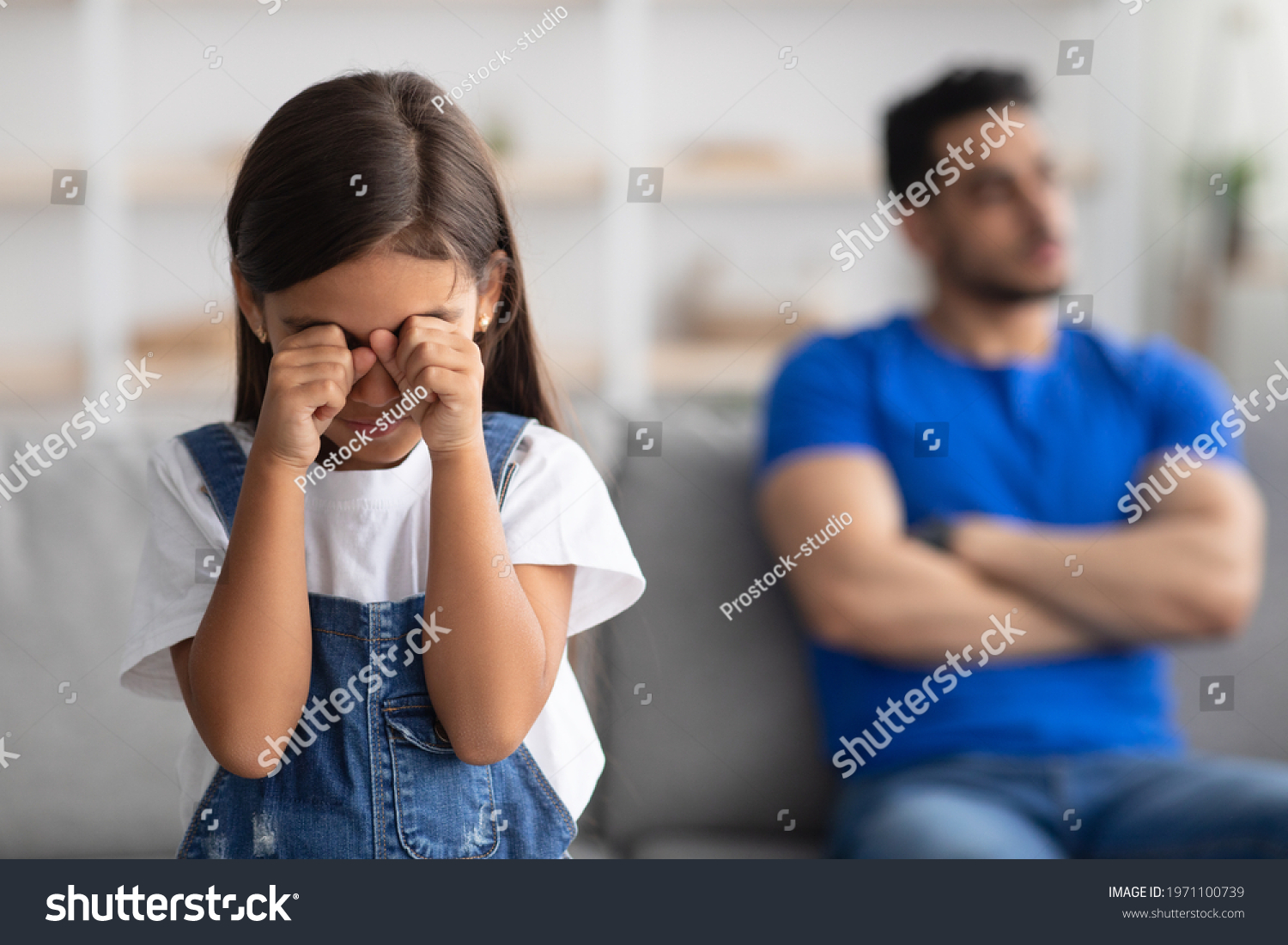 Family Relationship Problems. Selective focus on offended crying little kid girl wiping tears, young father sitting separate on couch in the blurred background, ignoring daughter. Generations gap #1971100739