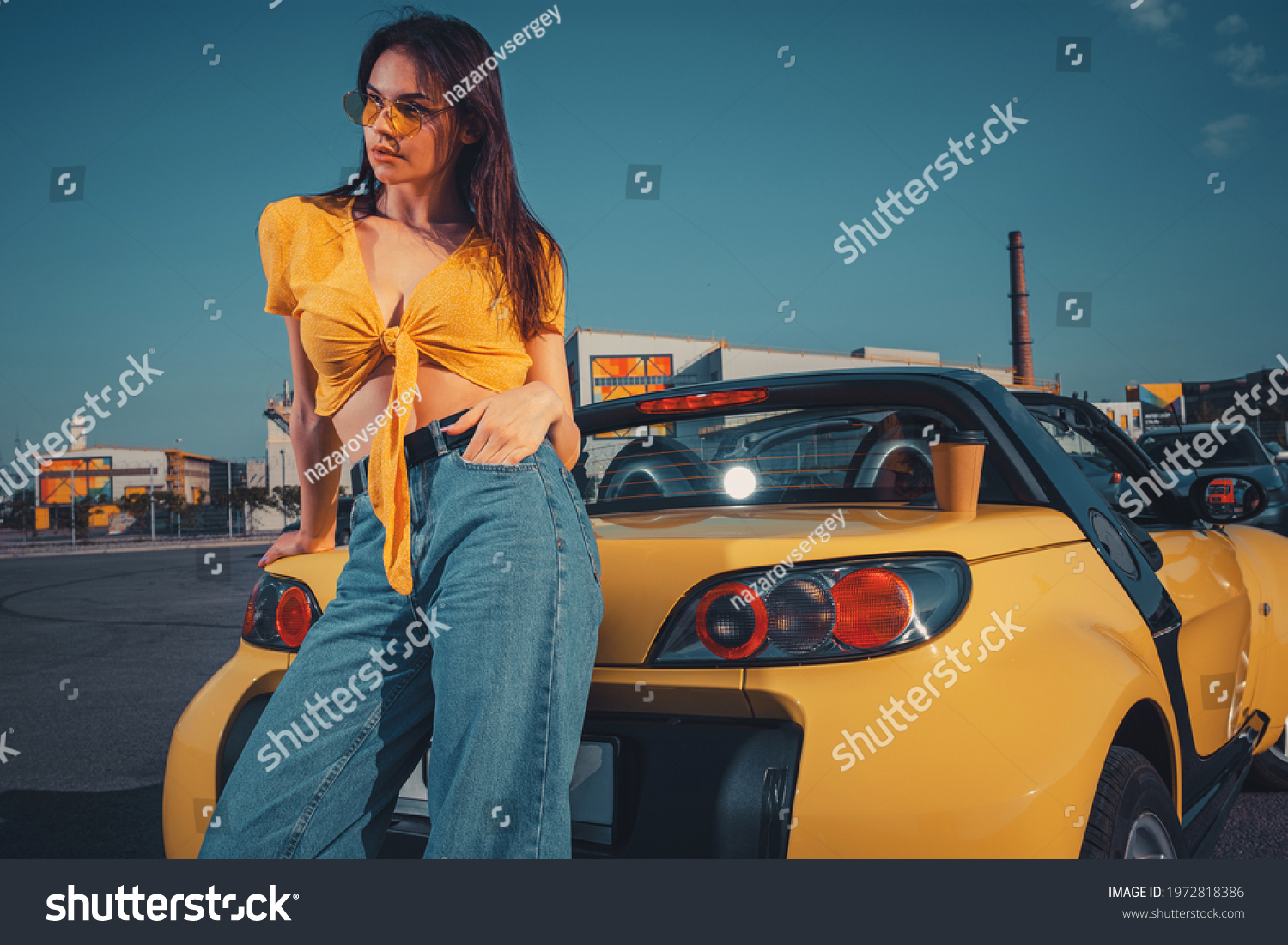 Lady in blue jeans, orange top and sunglasses is posing leaning on yellow car roadster with paper cup of coffee on trunk at parking lot. Close up #1972818386