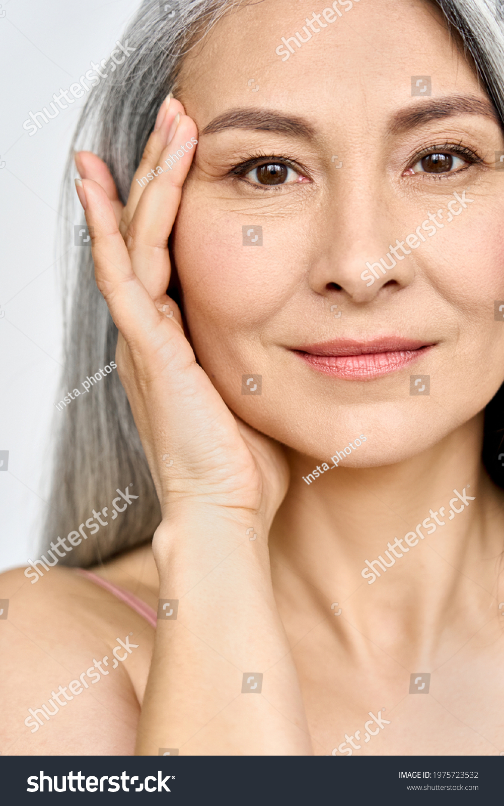 Vertical portrait of middle aged Asian woman's face with perfect skin. Older mature lady touching pampering face with hand. Advertising of cosmetology salon rejuvenating spa procedures skincare. #1975723532