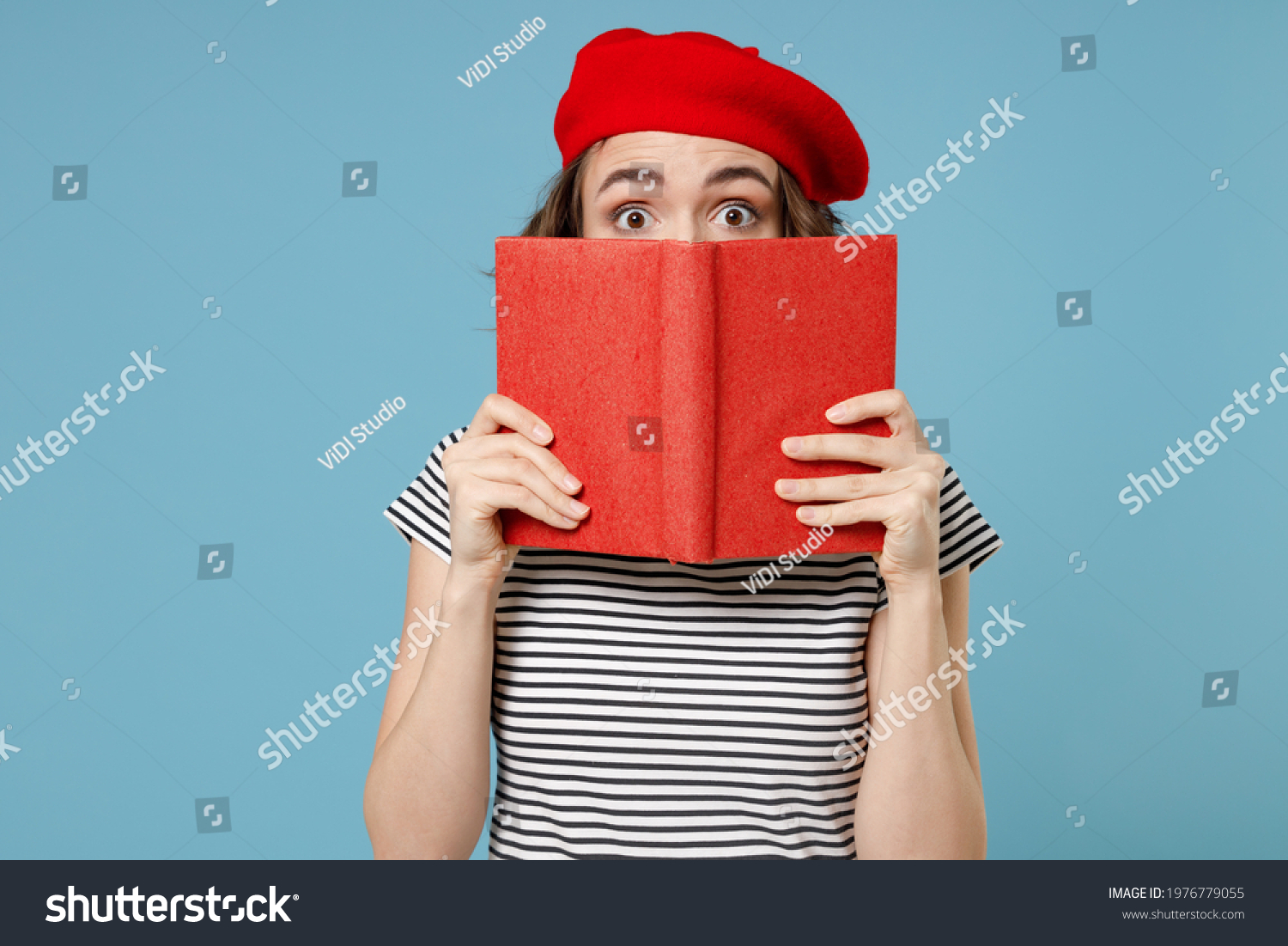 Young smart student woman 20s with short hairdo wearing french beret red hat striped t-shirt hiding cover face mouth with diary read book novel isolated on pastel blue color background studio portrait #1976779055