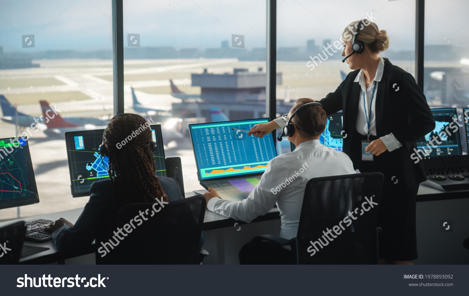 Female and Male Air Traffic Controllers with Headsets Talk in Airport Tower. Office Room is Full of Desktop Computer Displays with Navigation Screens, Airplane Departure and Arrival Data for the Team. #1978893092
