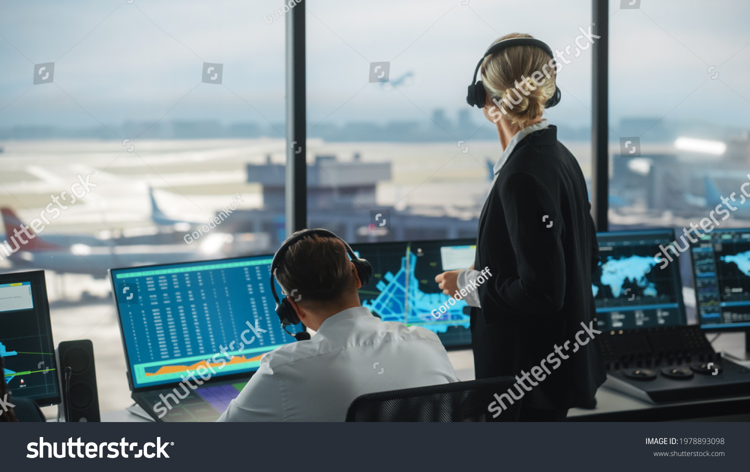 Female and Male Air Traffic Controllers with Headsets Talk in Airport Tower. Office Room is Full of Desktop Computer Displays with Navigation Screens, Airplane Departure and Arrival Data for the Team. #1978893098