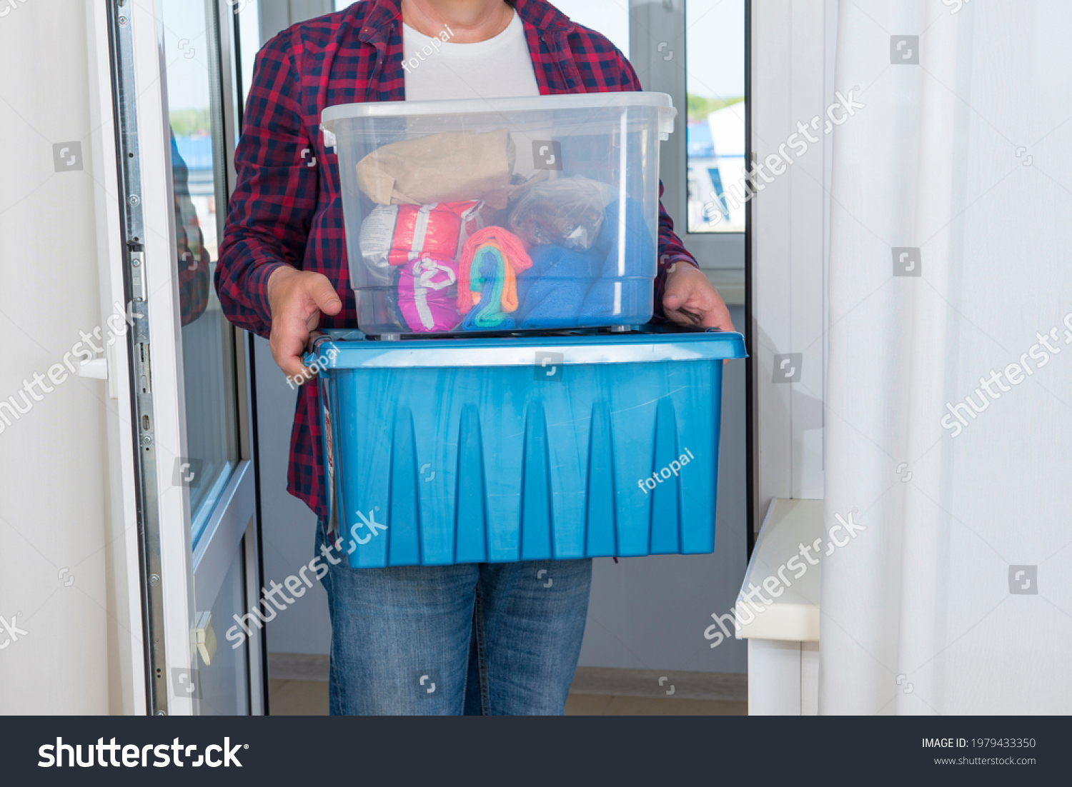 The man carries two plastic boxes with things, the upper transparent, the lower container is blue. #1979433350