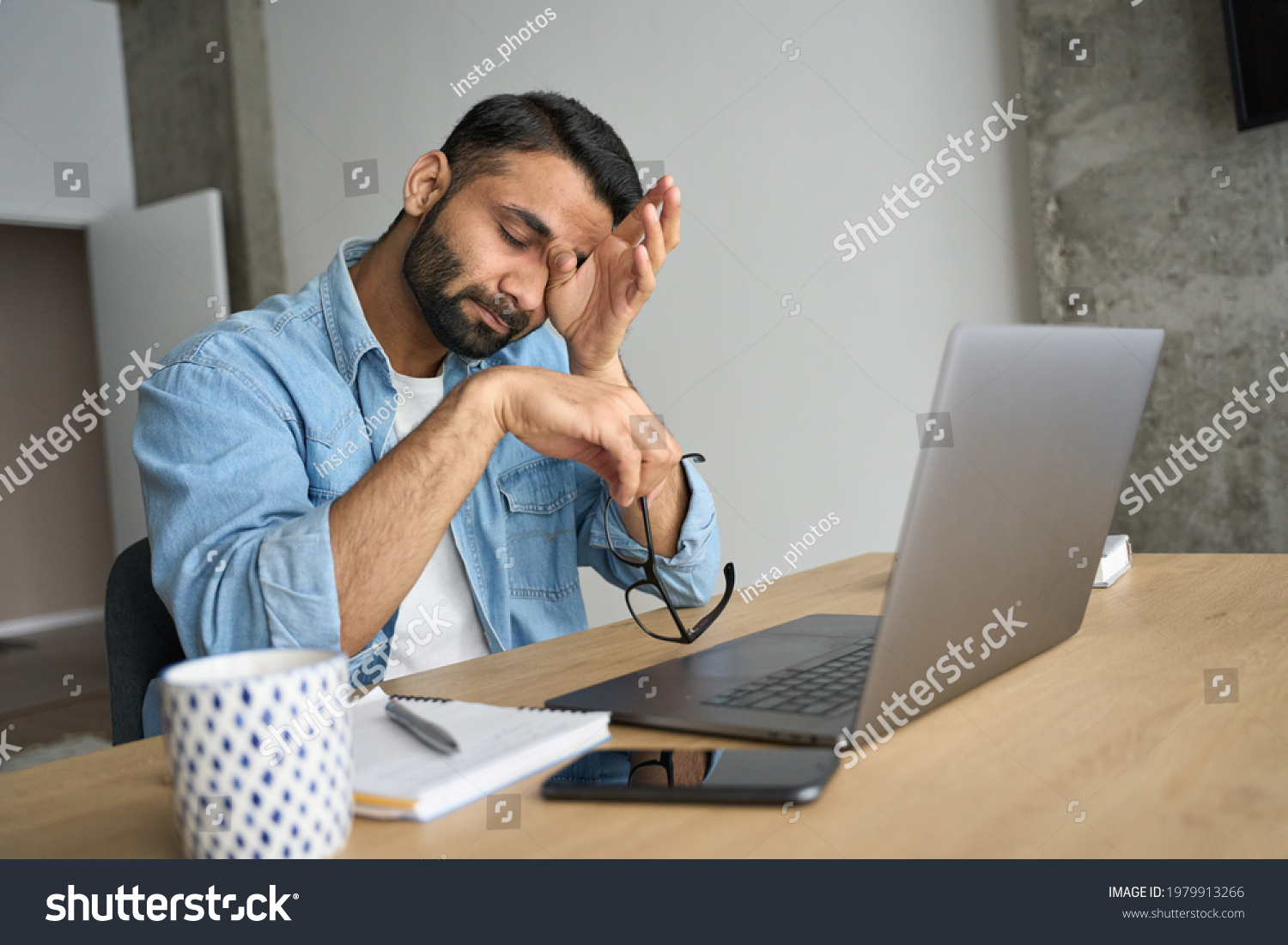 Young indian eastern tired exhausted business man rubbing eyes sitting in modern home office with laptop on desk. Overworked burnout academic Hispanic student with glasses in hand feeling eyestrain. #1979913266