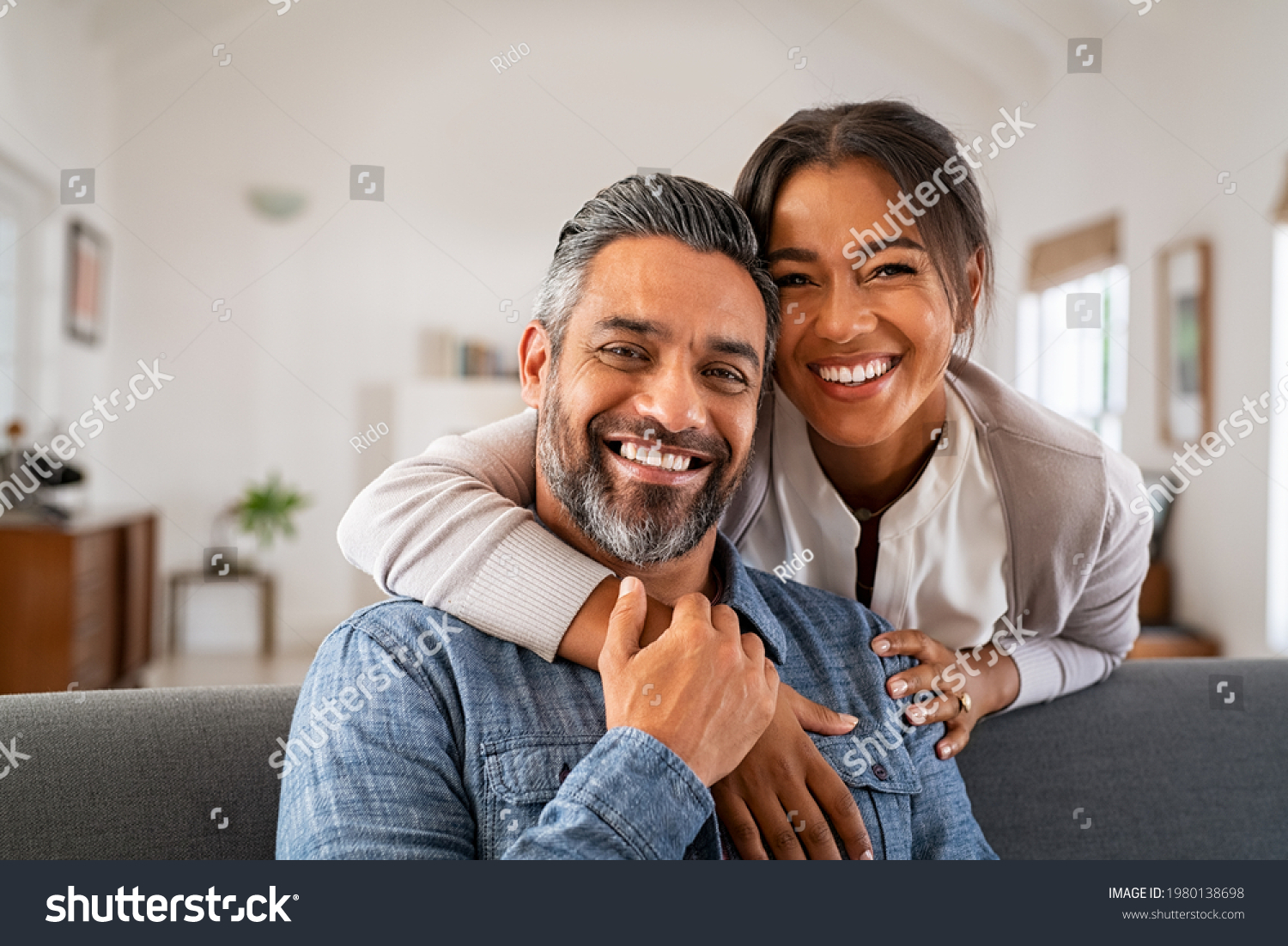 Portrait of multiethnic couple embracing and looking at camera sitting on sofa. Smiling african american woman hugging mid adult man sitting on couch from behind at home. Happy mixed race couple laugh #1980138698