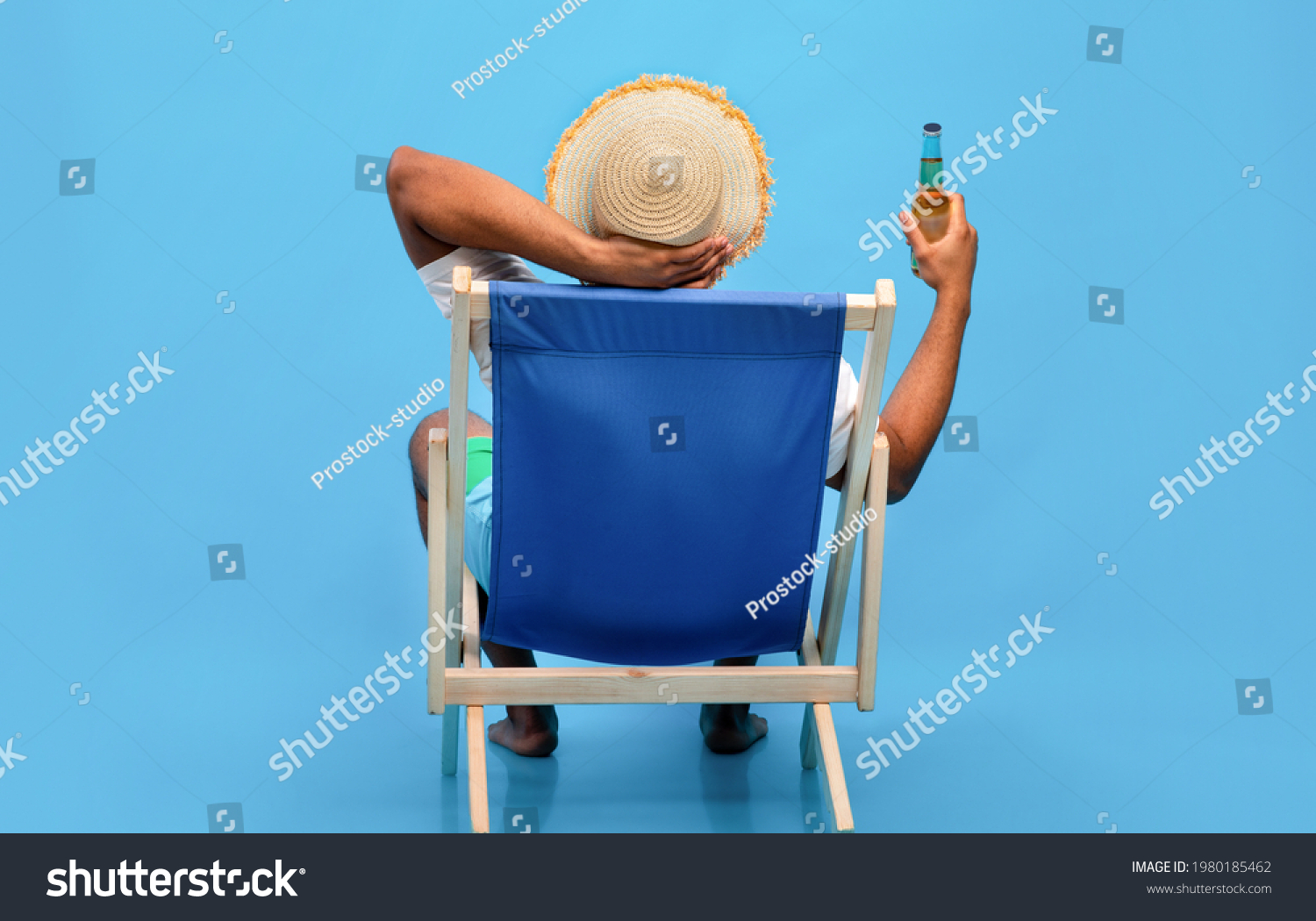 Back view of young black guy sitting in lounge chair with bottle of beer, enjoying summer vacation, sipping alcoholic beverage, sunbathing on blue studio background, full length #1980185462