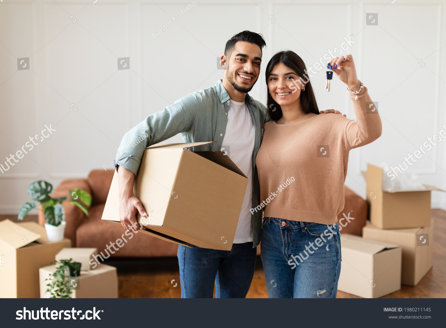 House Ownership. Young Couple Showing Keys And Holding Cardboard Box, Cheerful Guy And Lady Hugging After Moving In New Apartment Standing In Living Room. Insurance, Real Estate, Mortgage Concept #1980211145