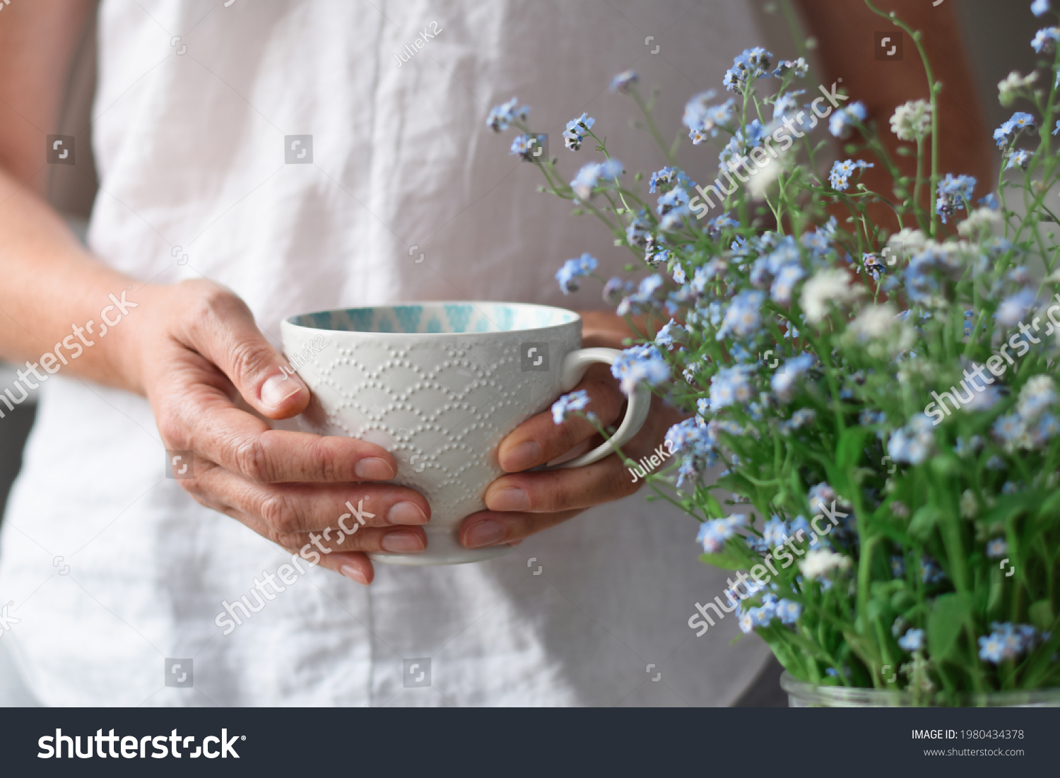 Woman's hands holding a white cup of tea or coffee beside blue flowers #1980434378