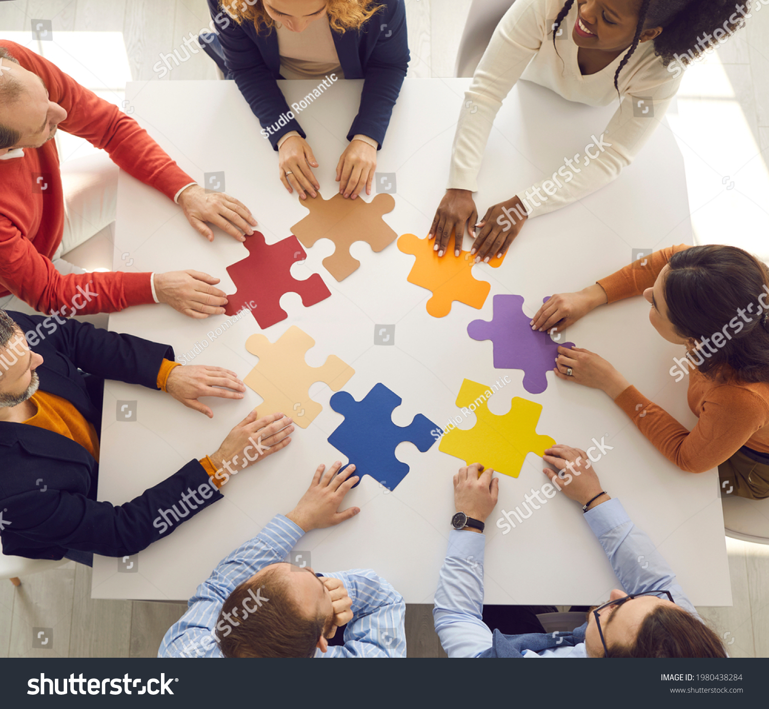 Team of young and senior employees sitting in circle around office table putting jigsaw puzzle together as metaphor for teamwork and looking for working business solutions. Square format, high angle #1980438284