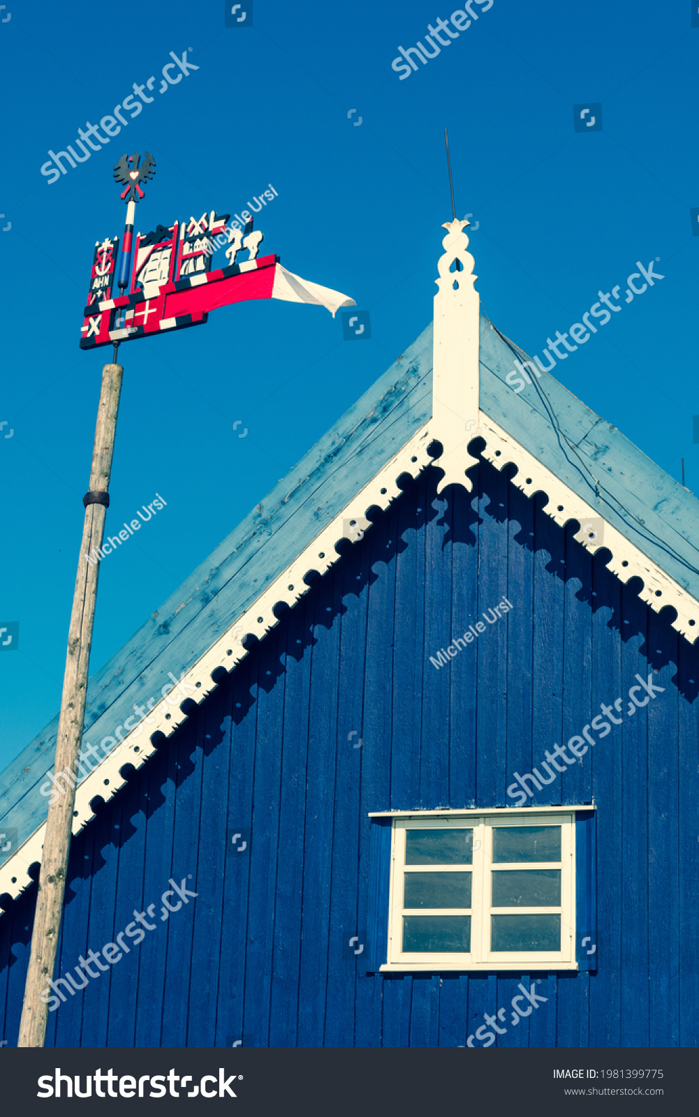 Beautiful colorful weathercock waving in the Curonian Spit in Nida fishermen's village, Lithuania, Europe with blue sky and roof of a wooden blue house with white window, vertical #1981399775