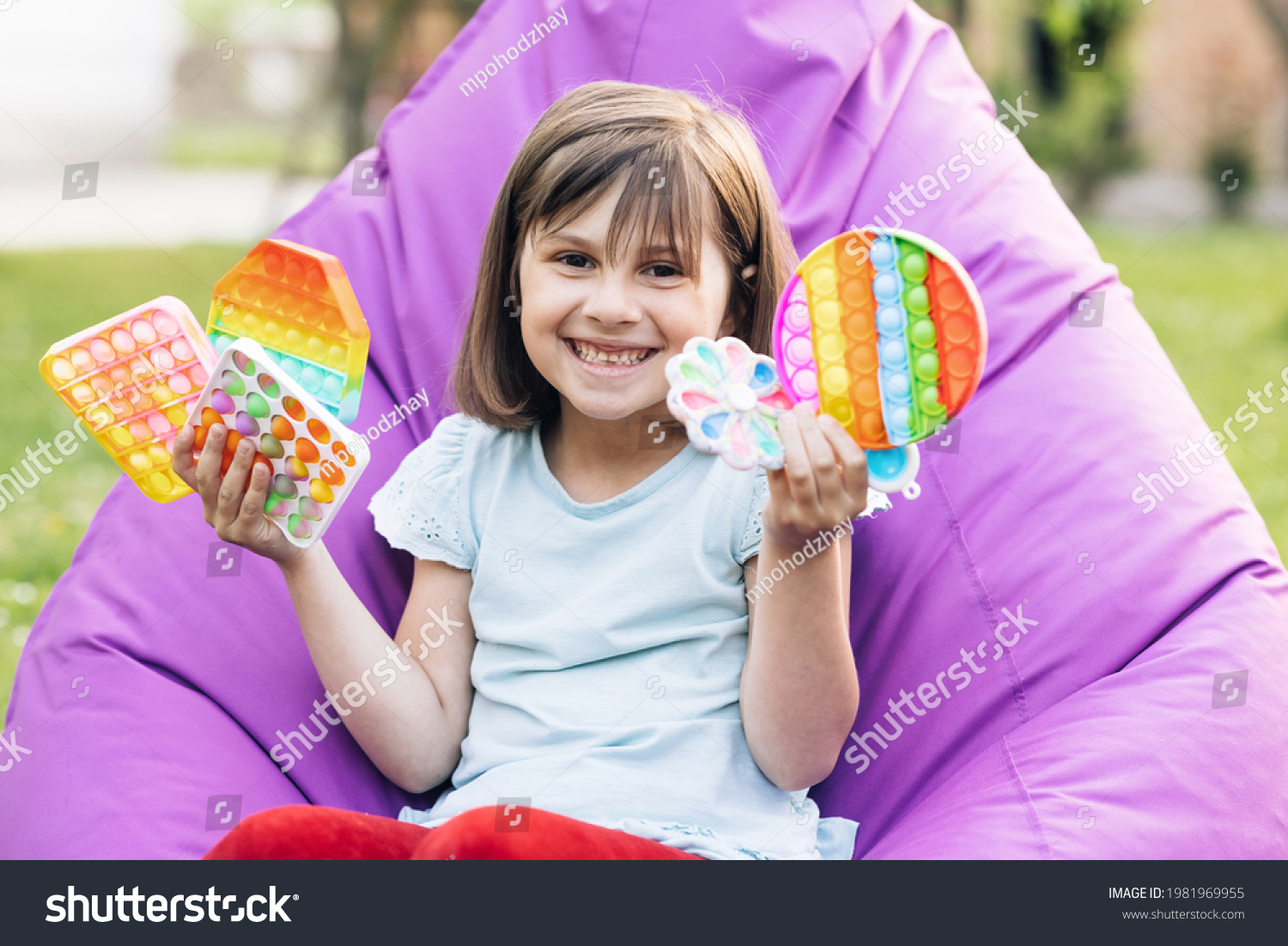 Portrait little girl with a modern popit toy. Colorful and bright pop it toy. Simple dimple. The child is sitting on a easy chair with colorful trendy antistress sensory toy pop it and simple dimple #1981969955