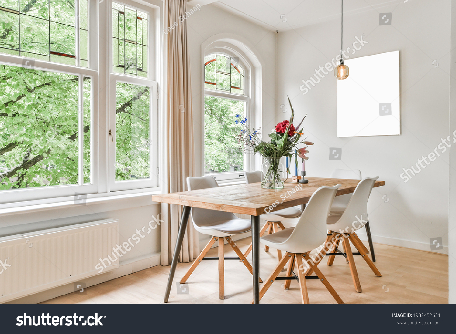 Table with flowers and chairs located near windows and abstract painting in light dining room at daytime #1982452631