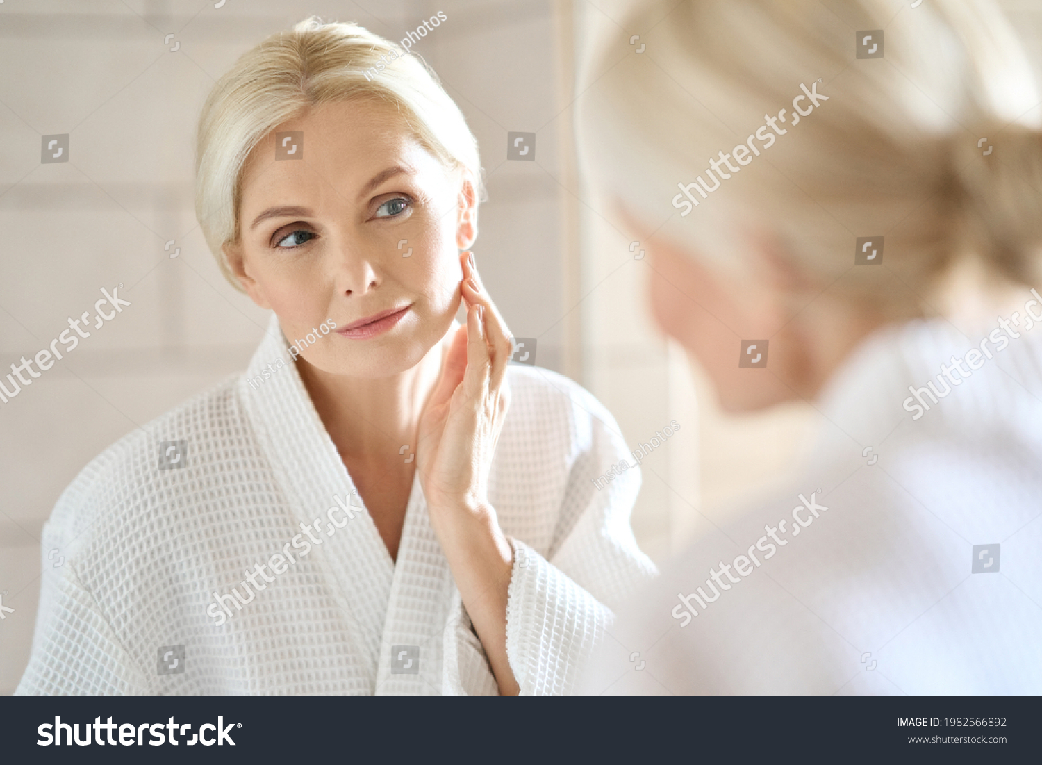 Headshot of gorgeous mid age adult 50 years old blonde woman standing in bathroom after shower touching face, looking at reflection in mirror doing morning beauty routine. Older skin care concept. #1982566892