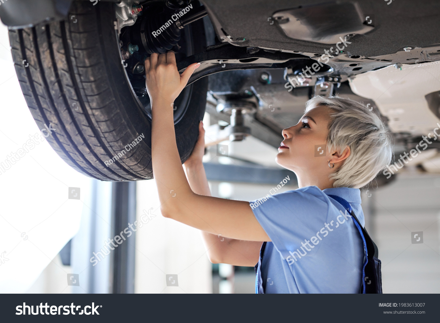 Caucasian female auto mechanic changing wheel tire in car in garage, side view. Beautiful young lady in overalls is concentrated on work, carefully adjusting repairing #1983613007