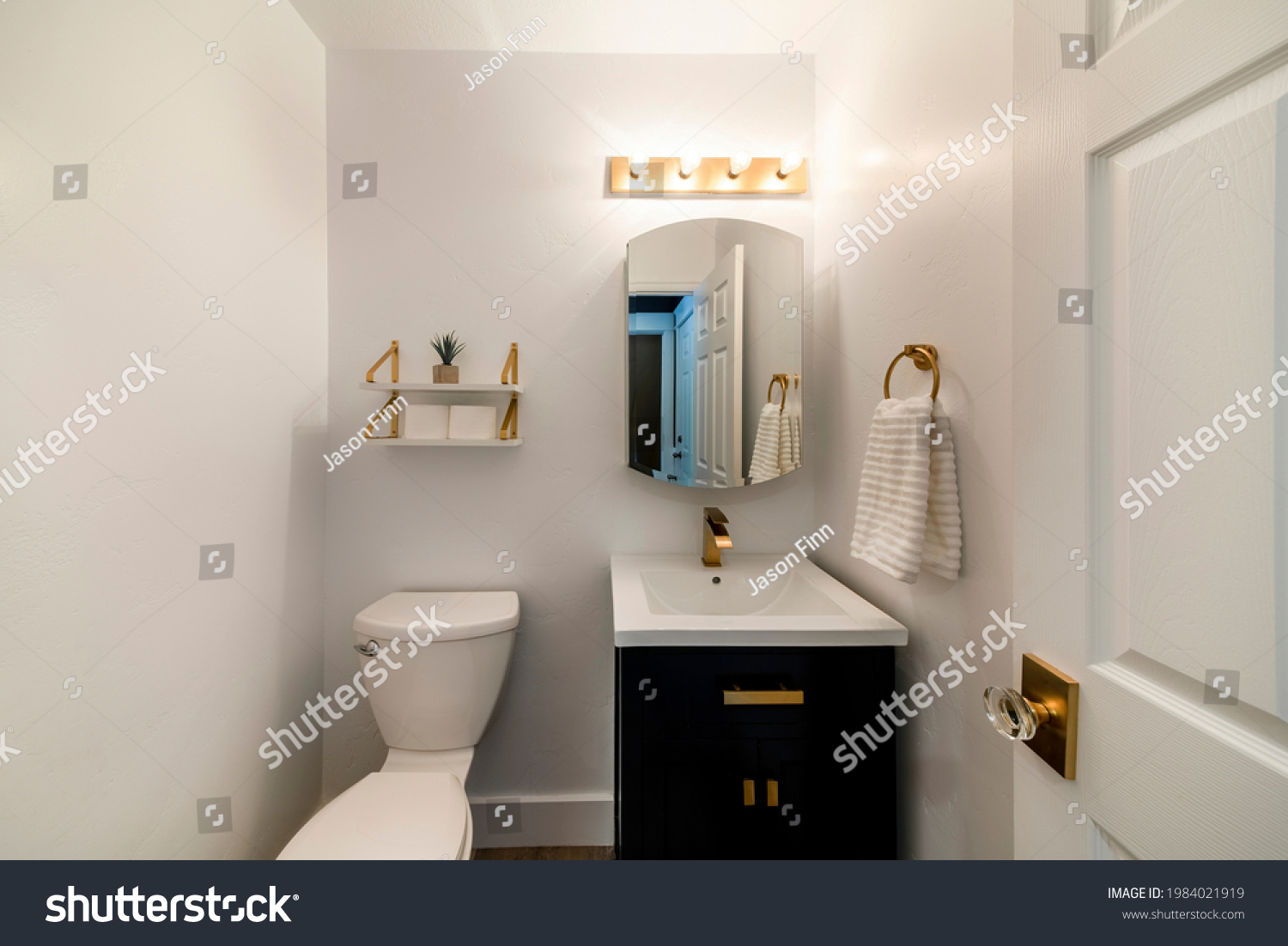Modern powder room design with matching gold fixtures. Single vanity sink with dark wood cabinet and round mirror beside the toilet bowl under the layered shelves with gold brackets. #1984021919