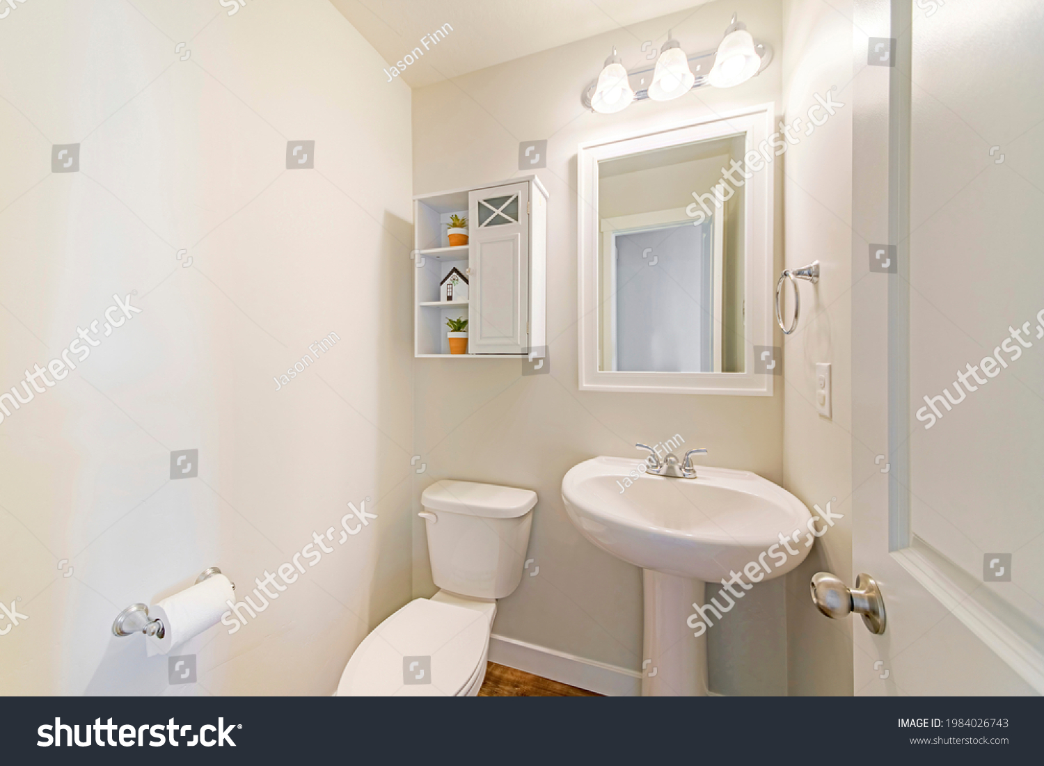 Interior of a white powder room with minimalist design. Toilet with ceramic bowl and sink with white wooden framed mirror beside the small white wooden cabinet and shelves with plants and house decor. #1984026743