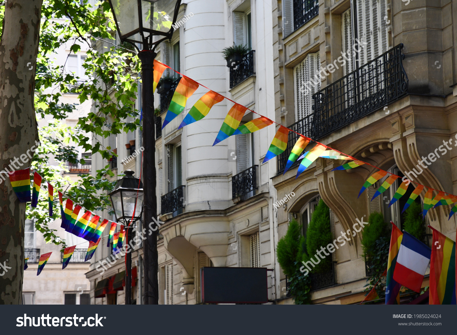 Decoration of triangle shape banners in colors of Lgbtq flags hanging between vintage lantern streetlights and ornate house with balconies. Gay pride parade symbols and French flag in Paris, France #1985024024